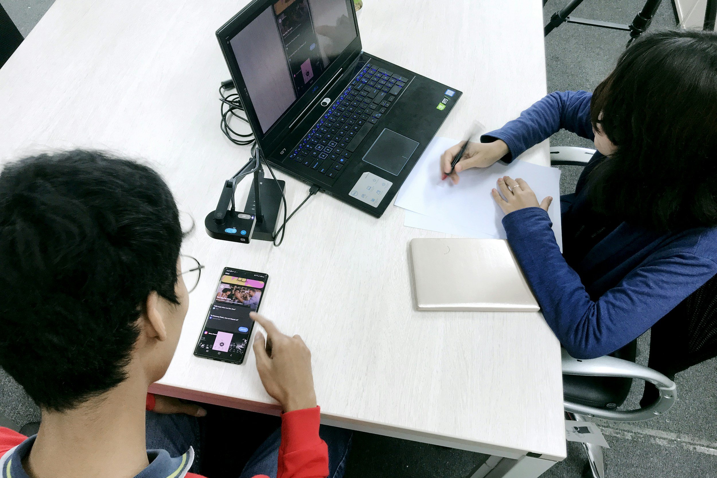 Two people sitting at a white conference table, one person using a smartphone and the other writing on paper near a laptop.