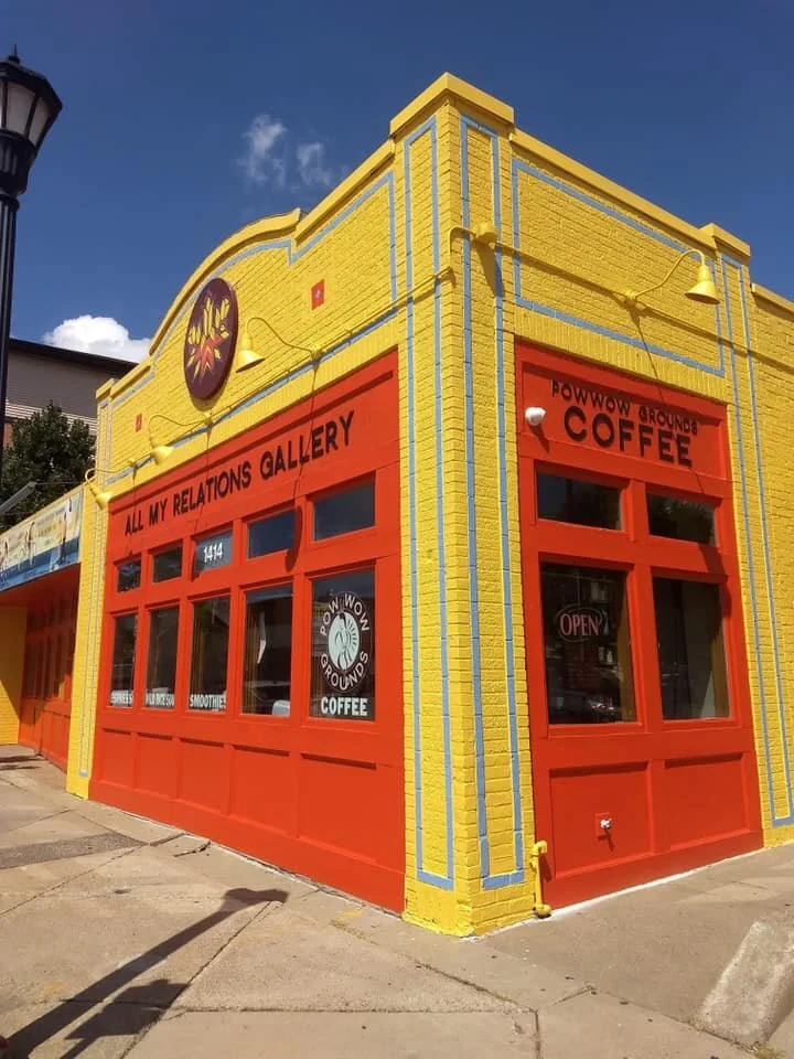 Colorful building with yellow and red walls, featuring signs for All My Relations Gallery and PowWow Grounds Coffee, with a blue sky overhead.