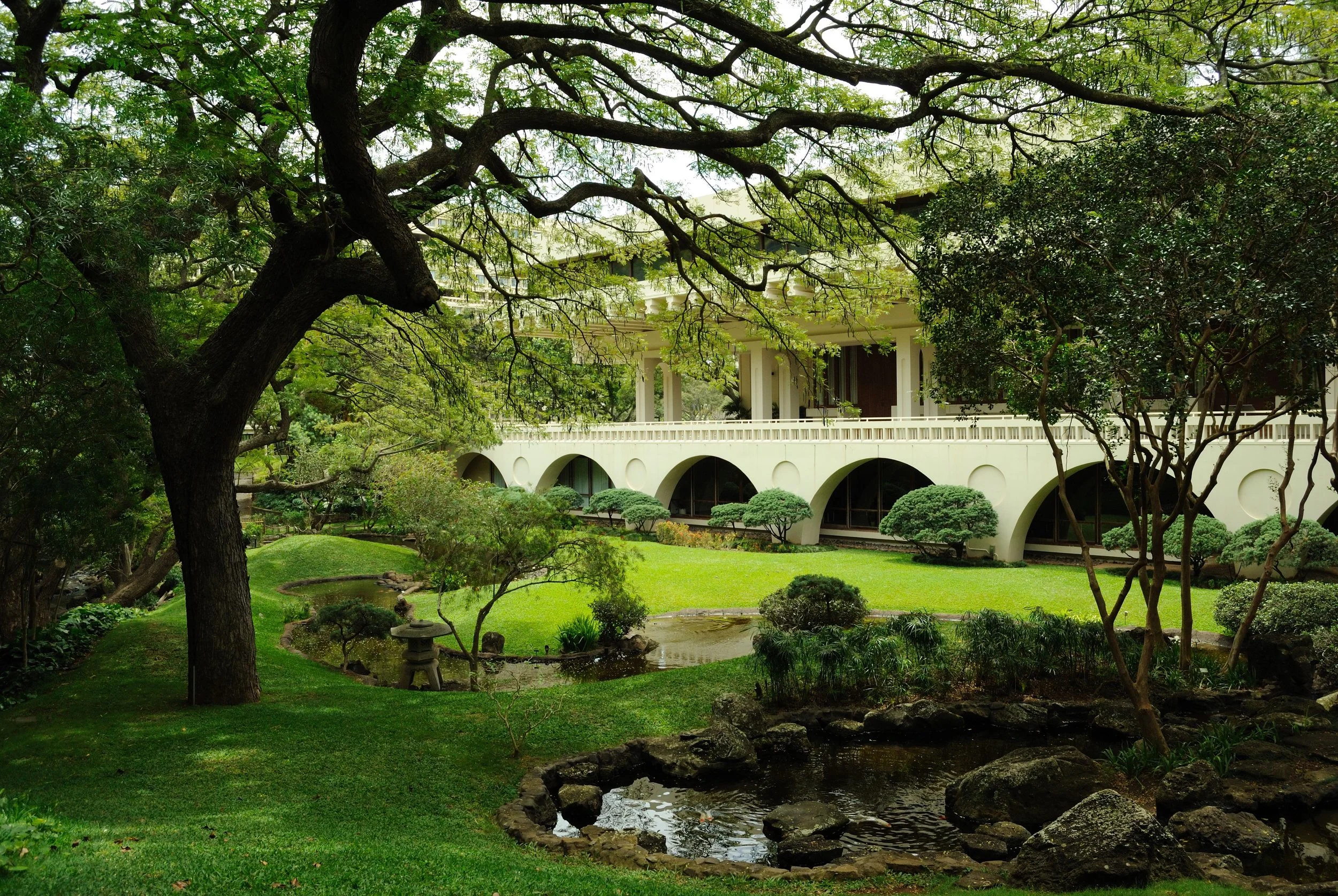 A large, modern building with a covered porch supported by columns, situated in a park-like setting with trees and green hills in the background.
