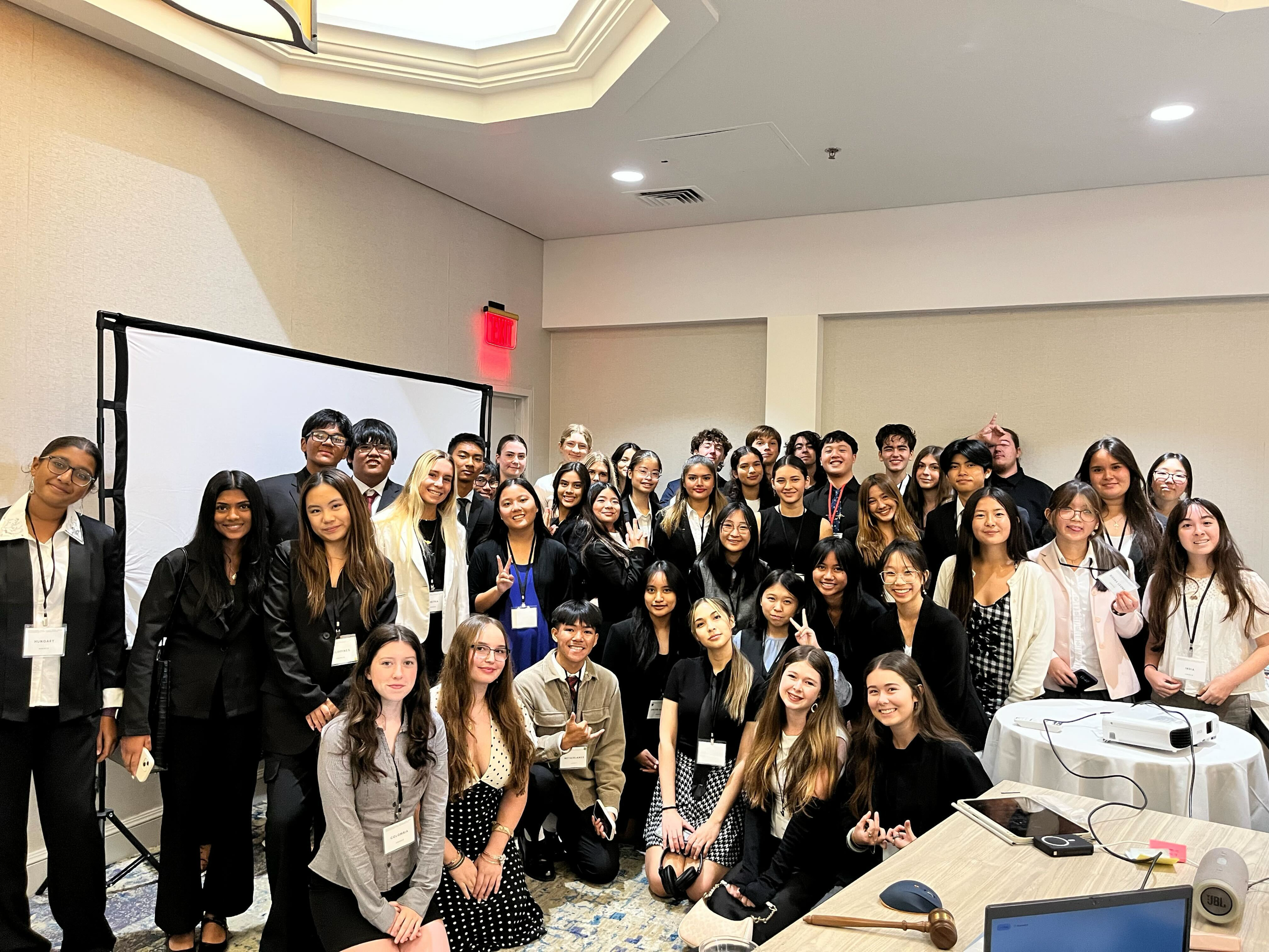 A large group of diverse young adults gathered in a conference room for a group photo. They are dressed in business casual attire, some wearing name tags, with a projector and laptop visible on a table in the foreground.