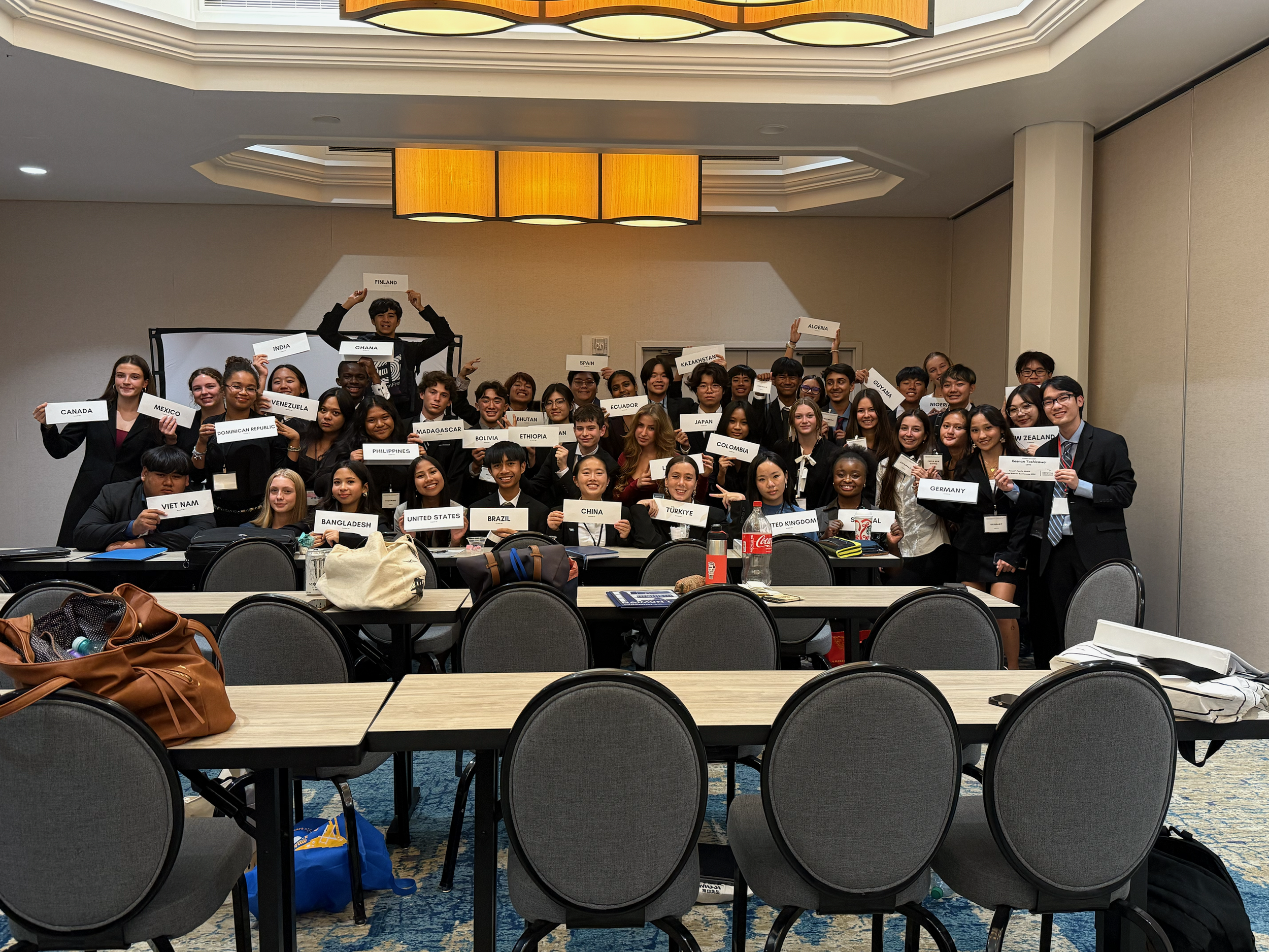 Group of diverse young adults at an indoor conference, holding signs with country names, with tables and empty chairs in the foreground.