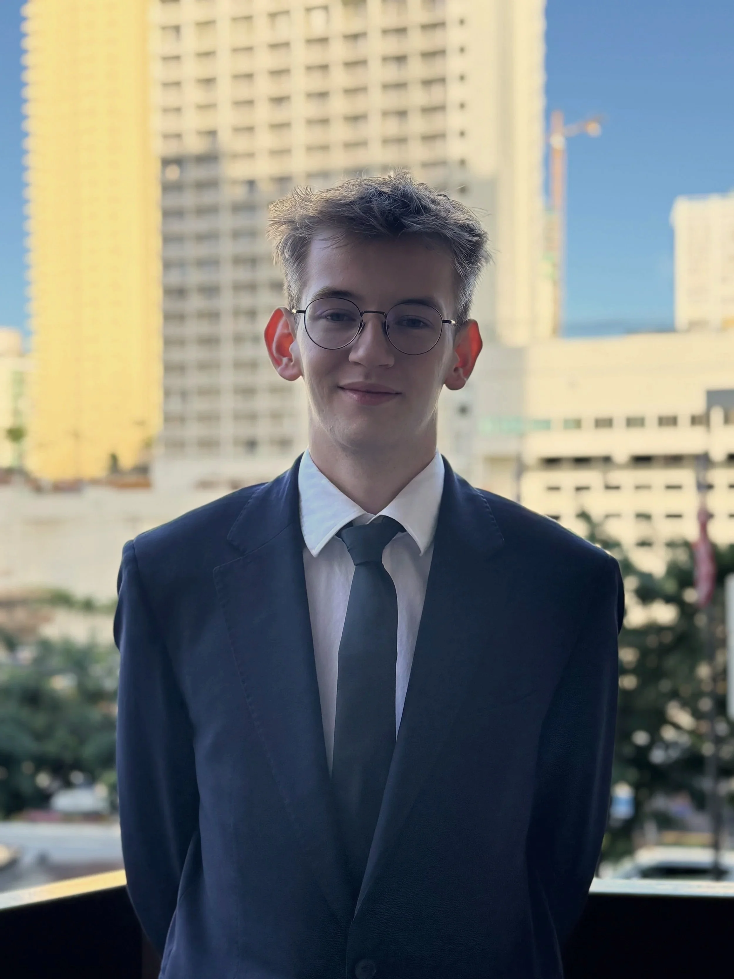A young man with light brown hair, glasses, and a slight smile, wearing a navy blue suit, white shirt, and black tie, standing outdoors with tall yellow and white buildings and a blue sky in the background.