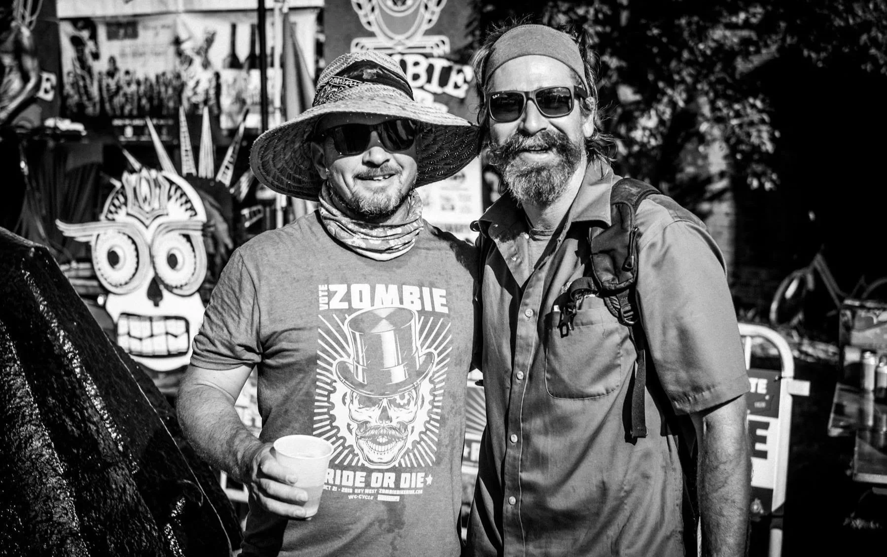Two men standing close together outdoors, smiling at the camera with trees and decorative banners in the background. One man wears a wide-brimmed hat, sunglasses, and a T-shirt with a skull graphic and text. The other man sports a headband, sunglasses, a beard, and a backpack.