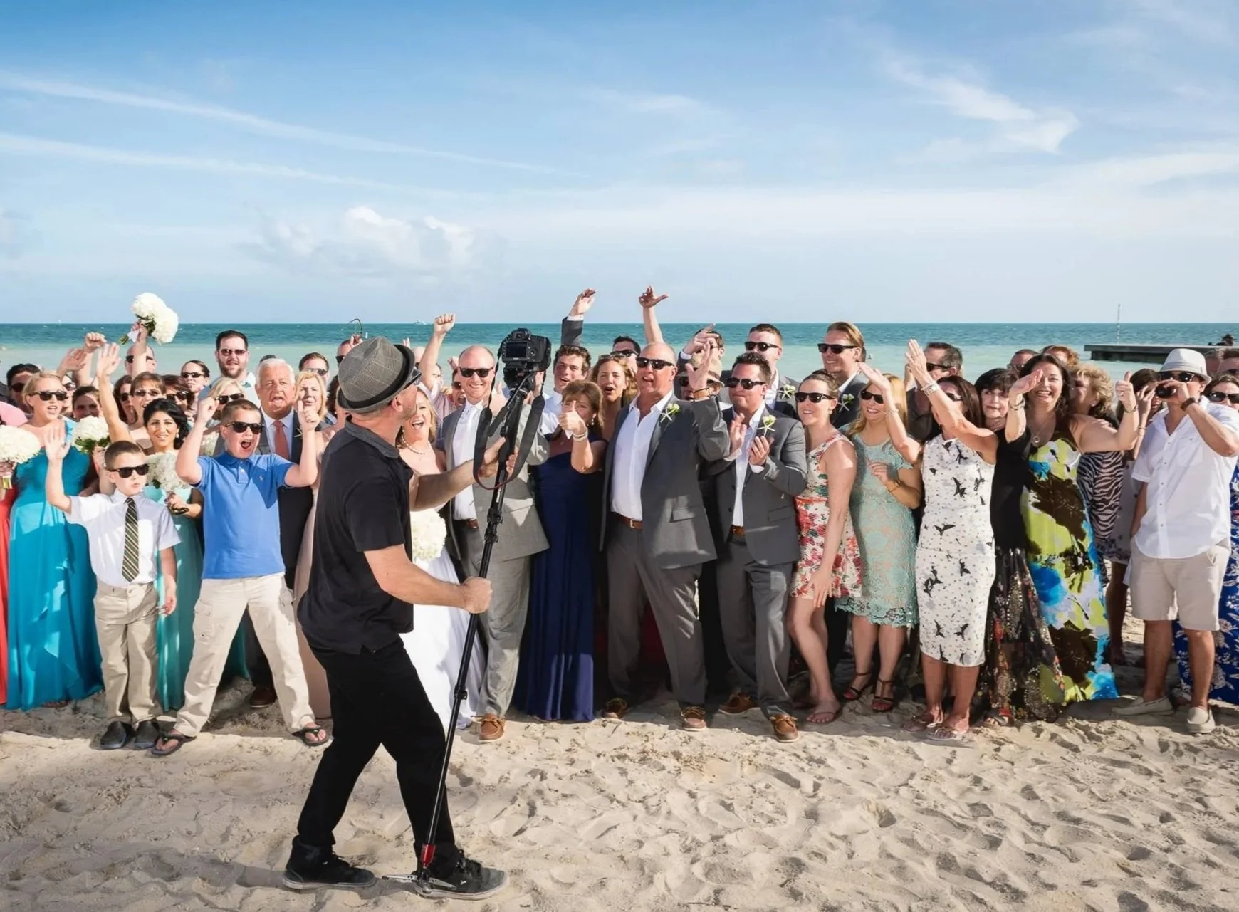 A large group of people celebrating at the beach, likely for a wedding or special event, with the ocean and sky in the background. The group is dressed in formal and semi-formal clothing, and someone is filming or taking photos of the gathering.