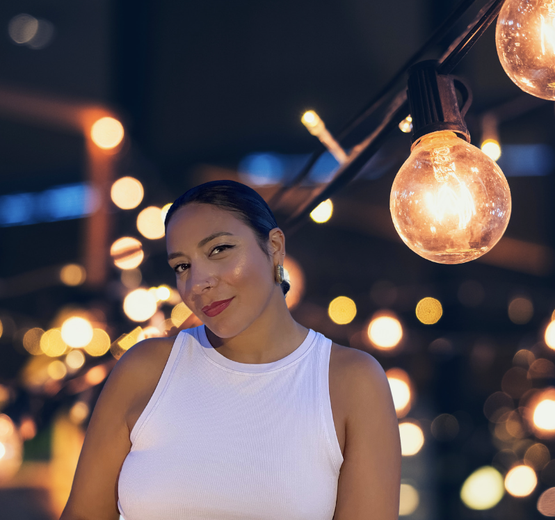 A woman with dark hair in a bun, wearing a white sleeveless top, smiling with a night cityscape background with string lights and glowing bulbs.