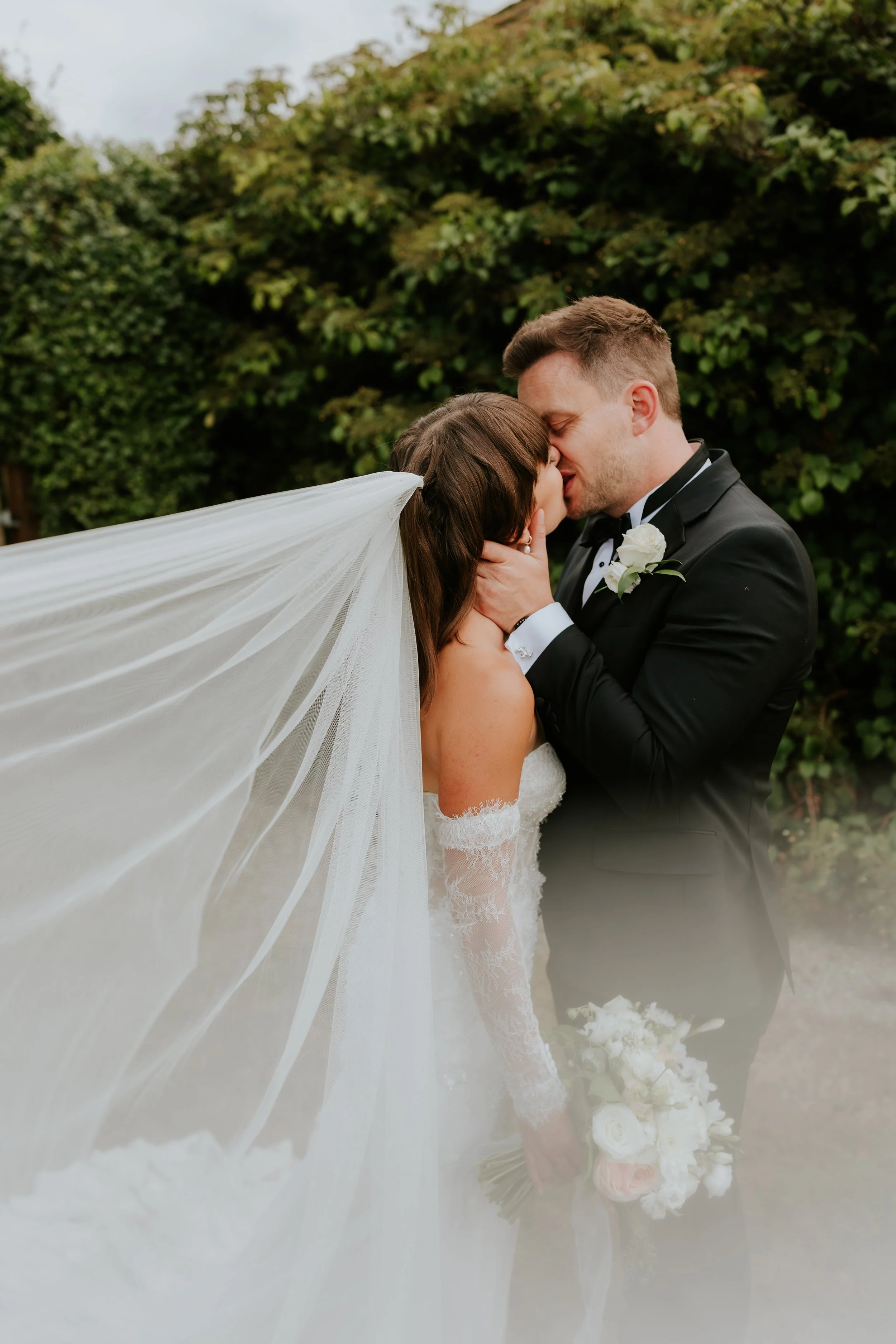 A newlywed couple shares a kiss outdoors, with the bride in a white wedding gown and veil, and the groom in a black tuxedo with a white boutonniere, holding a bouquet of white flowers.
