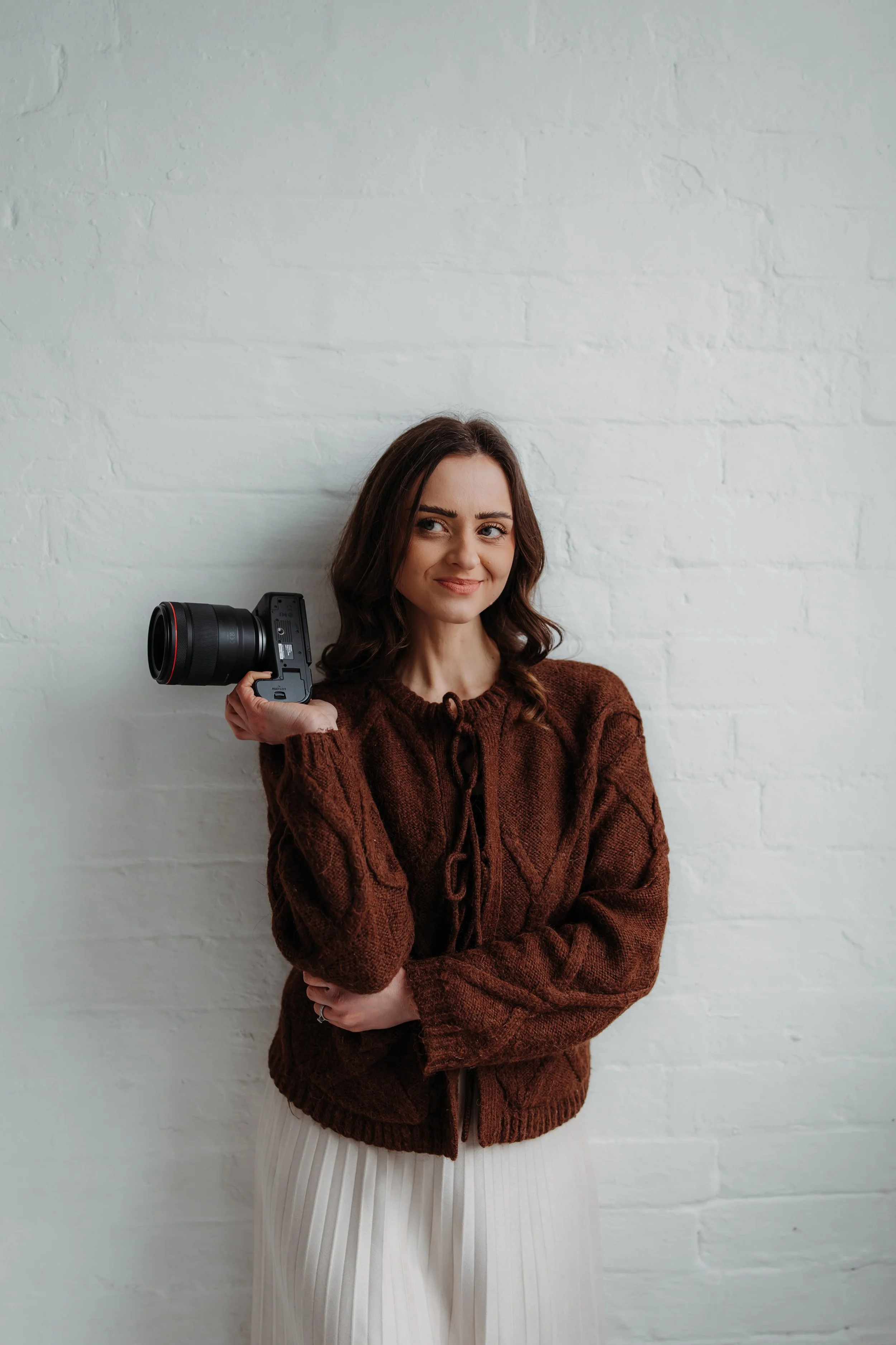 A woman with brown hair standing against a white brick wall, holding a camera on her shoulder, wearing a brown sweater and white pants.