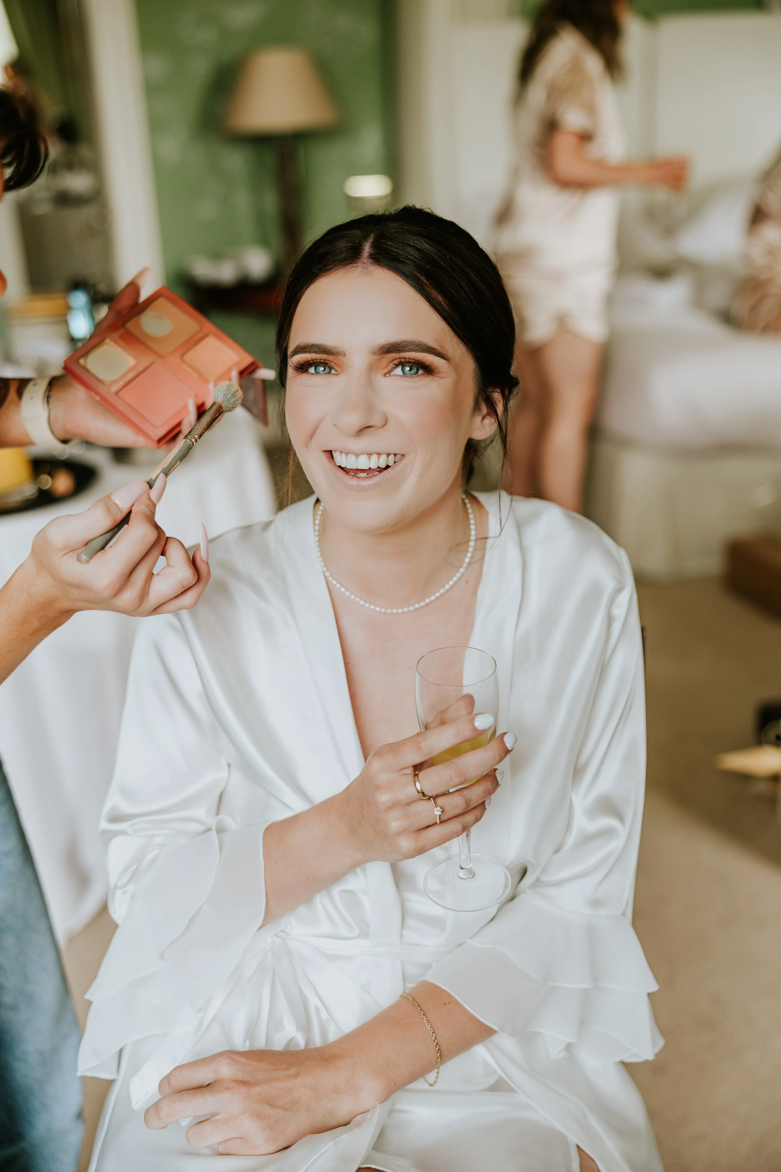Bridal woman getting her makeup done, holding a glass of champagne, smiling, wearing a white satin robe, pearl necklace, and ring, in a room with green walls and other women in the background.