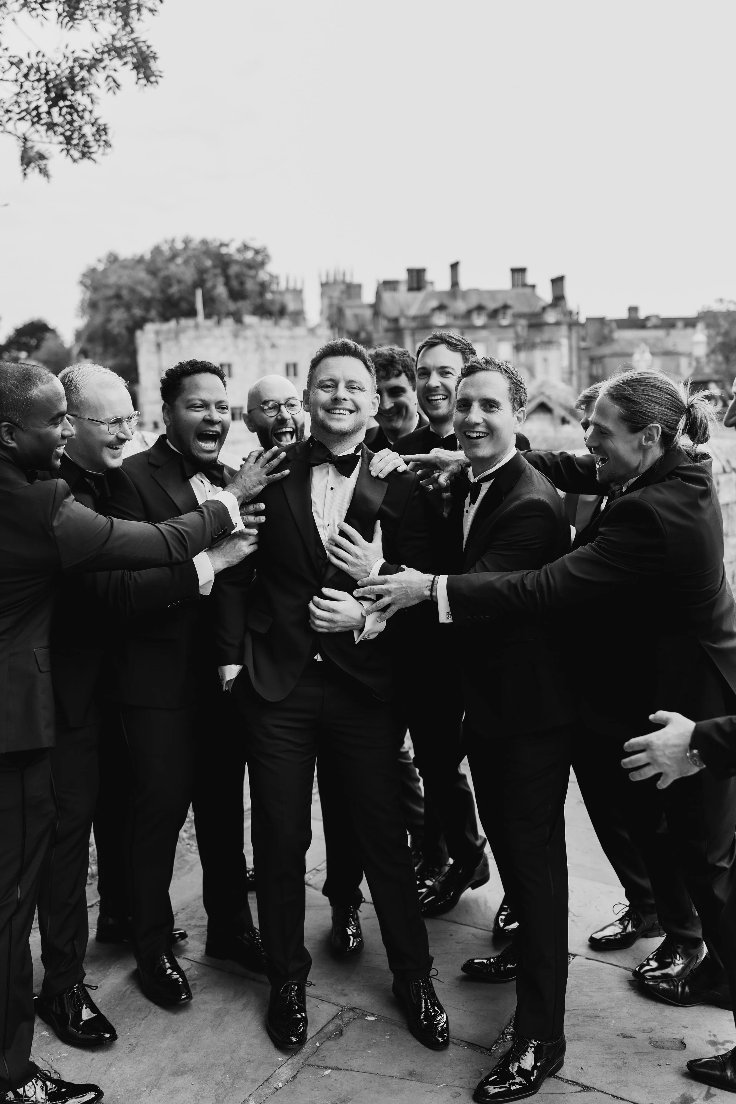 Black and white photo of a group of men in tuxedos, laughing and celebrating outdoors, with some touching or reaching toward a man in the center.