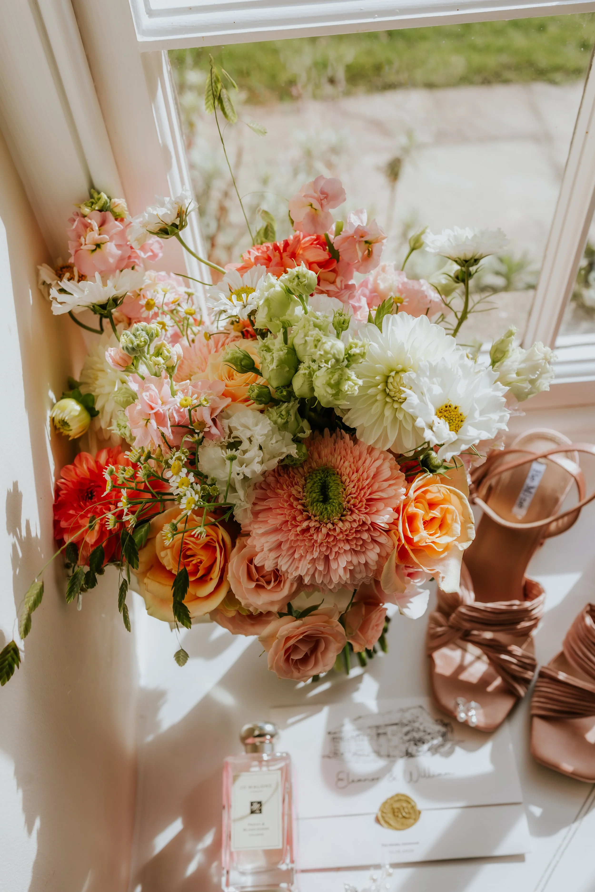 A bouquet of pink, white, and cream flowers on a windowsill, with a pair of pink high-heeled shoes and a perfume bottle nearby.