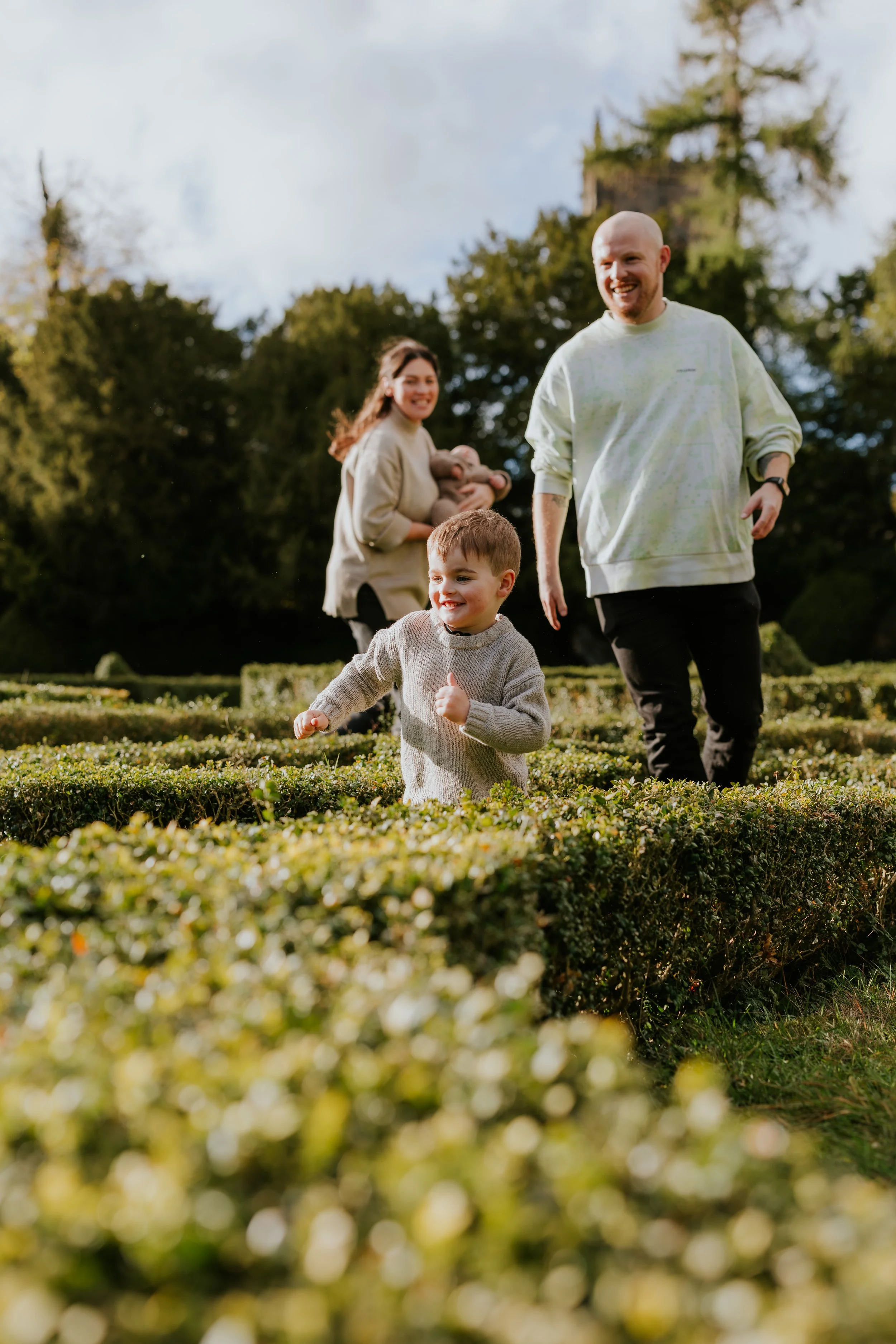 A family of three, including a young boy, a woman, and a man, running and smiling in a garden with neatly trimmed bushes, trees, and a stone tower in the background on a sunny day.