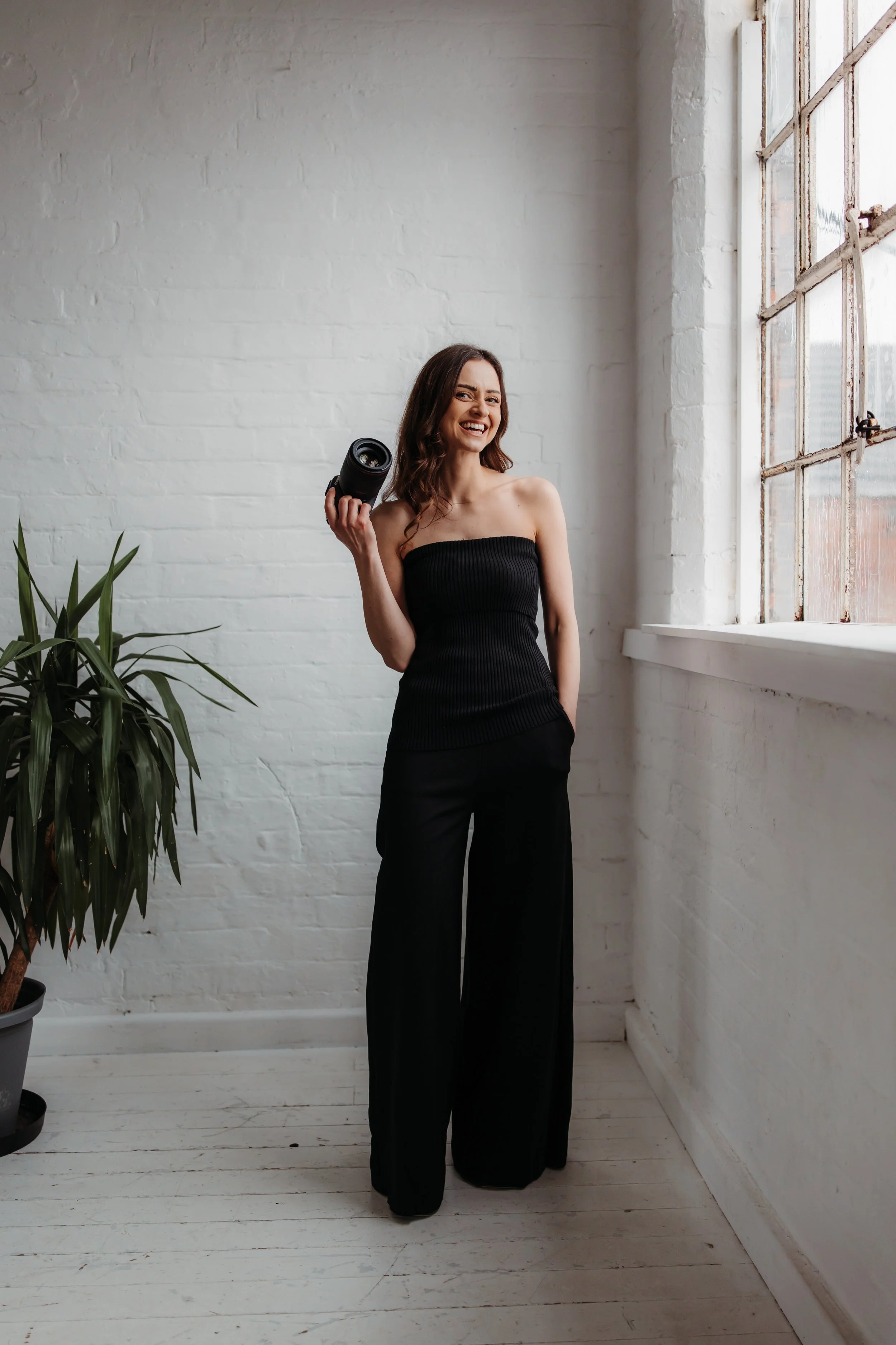 A woman with brown hair standing against a white brick wall, holding a camera on her shoulder, wearing a brown sweater and white pants.