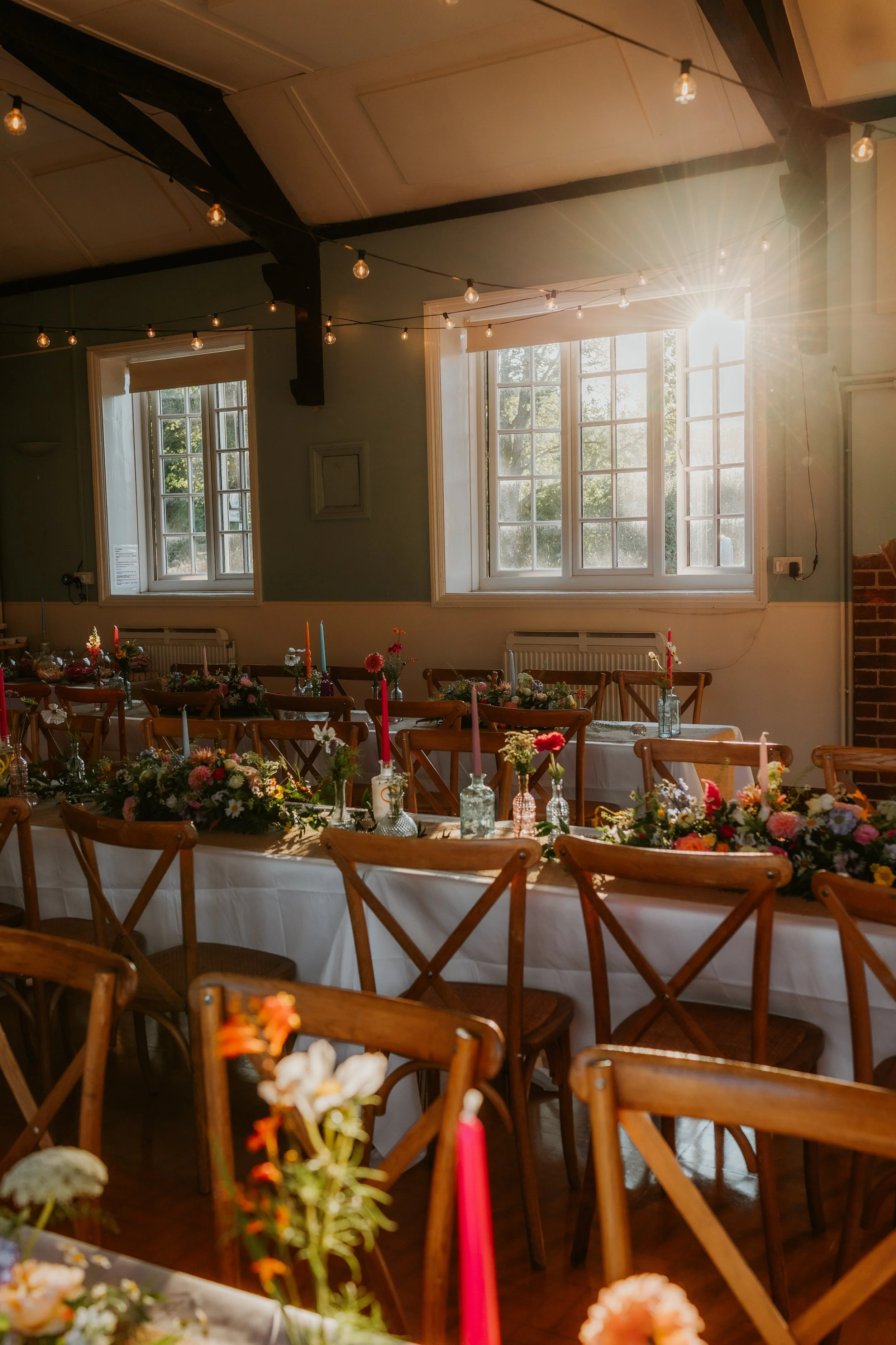 Sunlight streaming through large windows illuminating a decorated dining room with wooden chairs, floral arrangements, and candles on a long table.