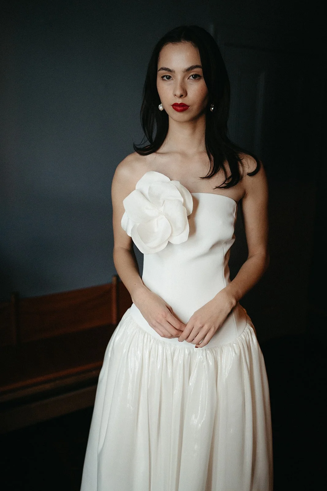 A woman in a strapless white gown with a large floral embellishment on her shoulder stands against a dark background.