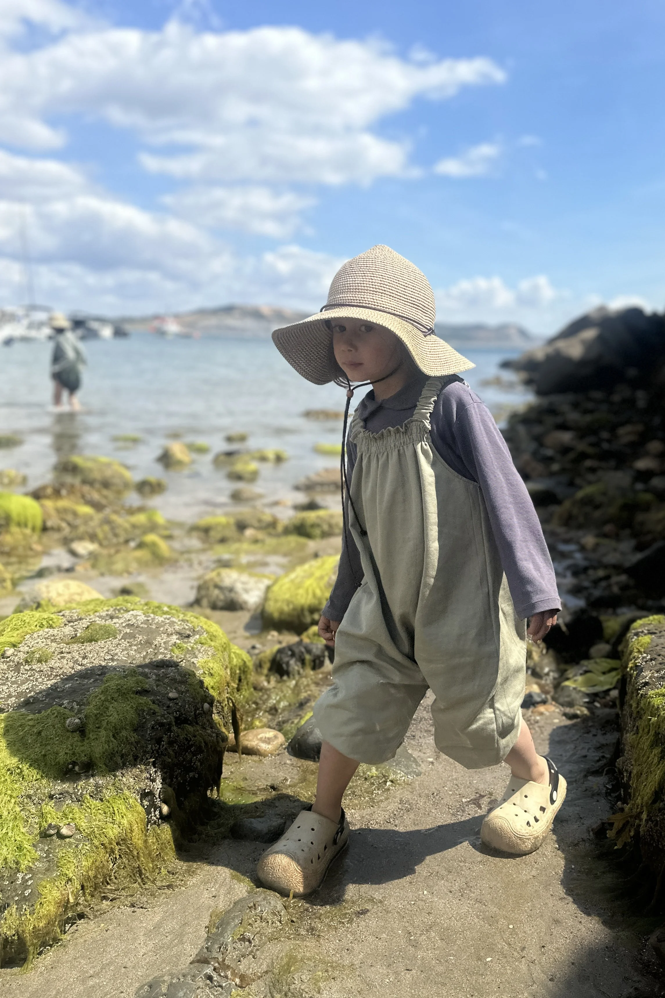 child wearing organic cotton denim light sage green pullover dungarees layered with purple top, sun hat, standing in the sea