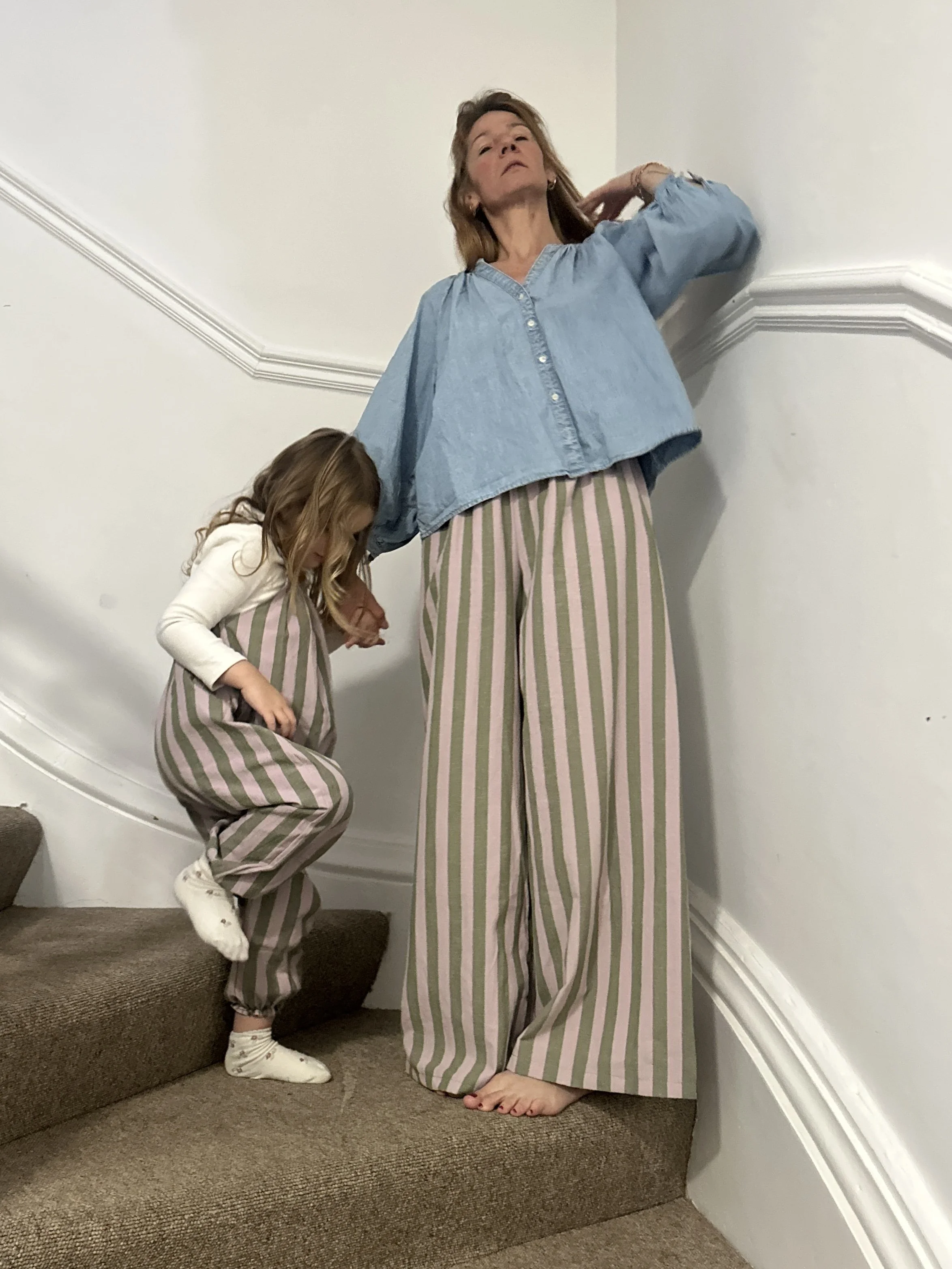 mum wearing organic cotton linen wide leg trousers stripes olive green and lilac, with denim light blue blouse standing on the stairs, next to a child wearing dungarees made of same fabric