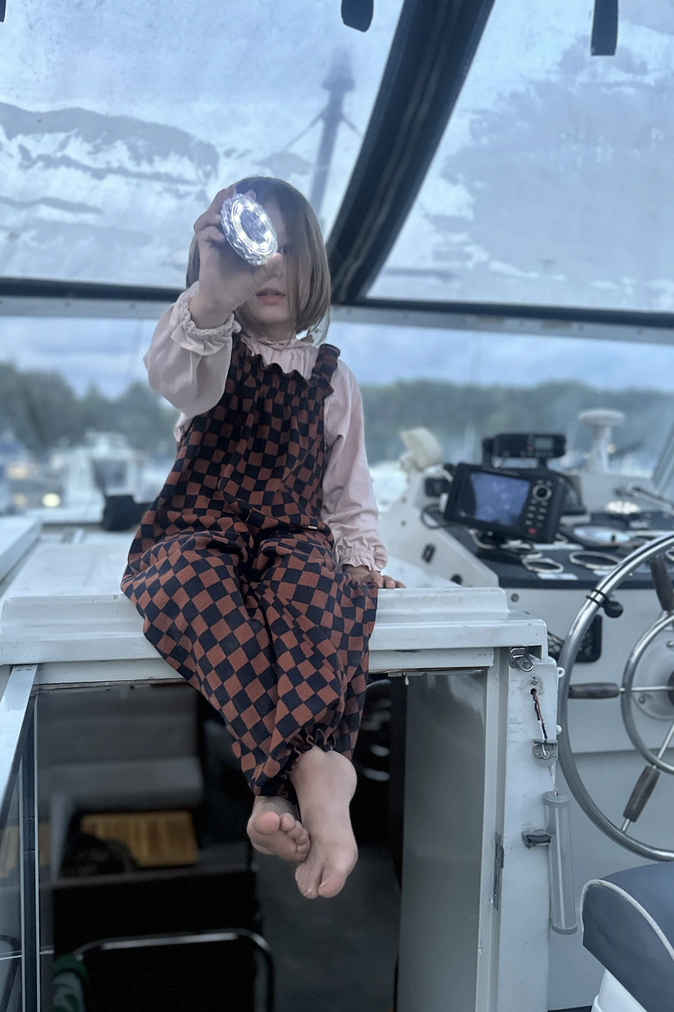 child wearing pullover dungarees in checkerboard navy and rust colour, sitting on a boat at marina