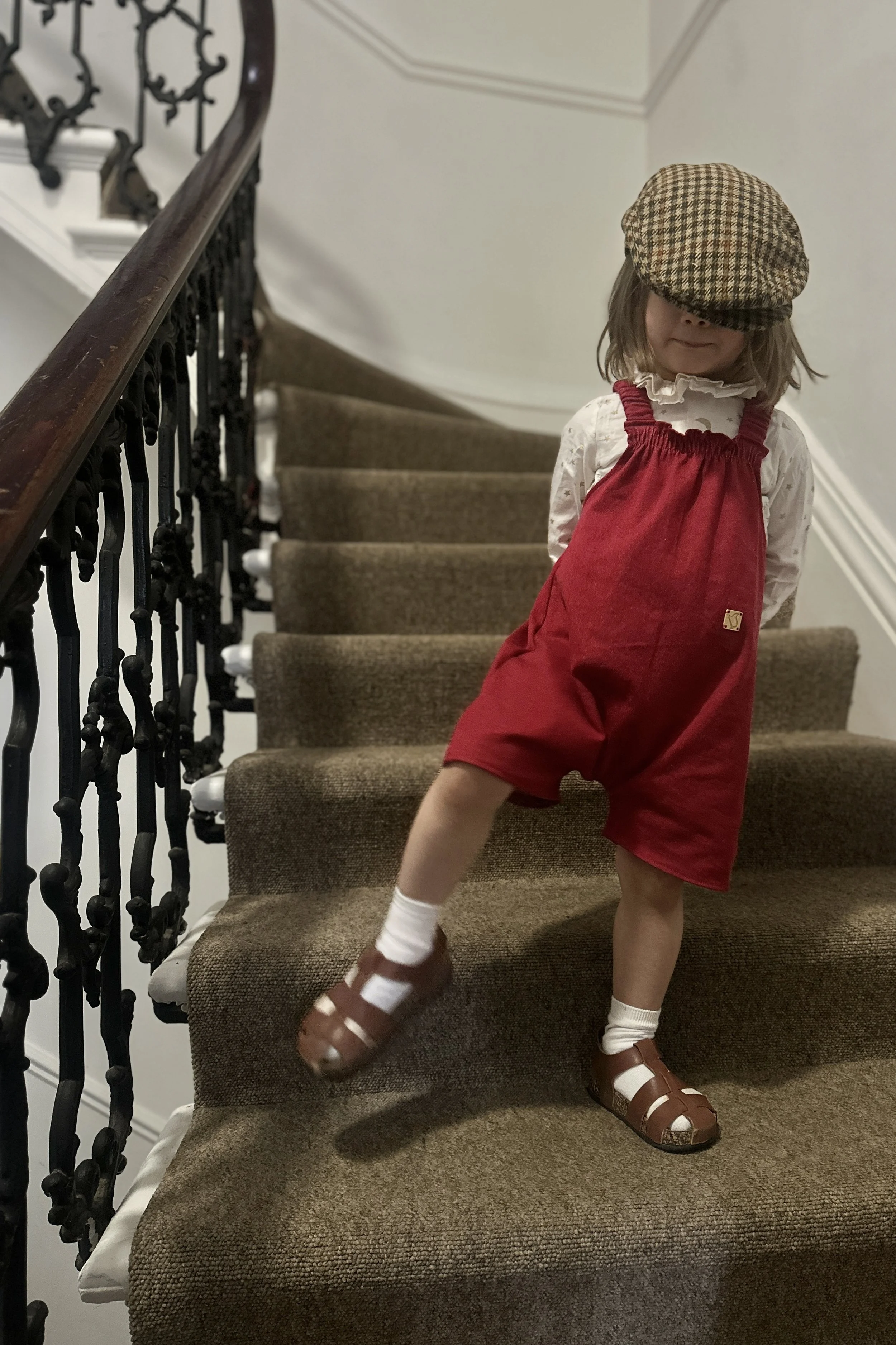 child wearing organic cotton denim red short dungarees over white blouse with golden stars with brown sandals tartan hat, standing on staircase