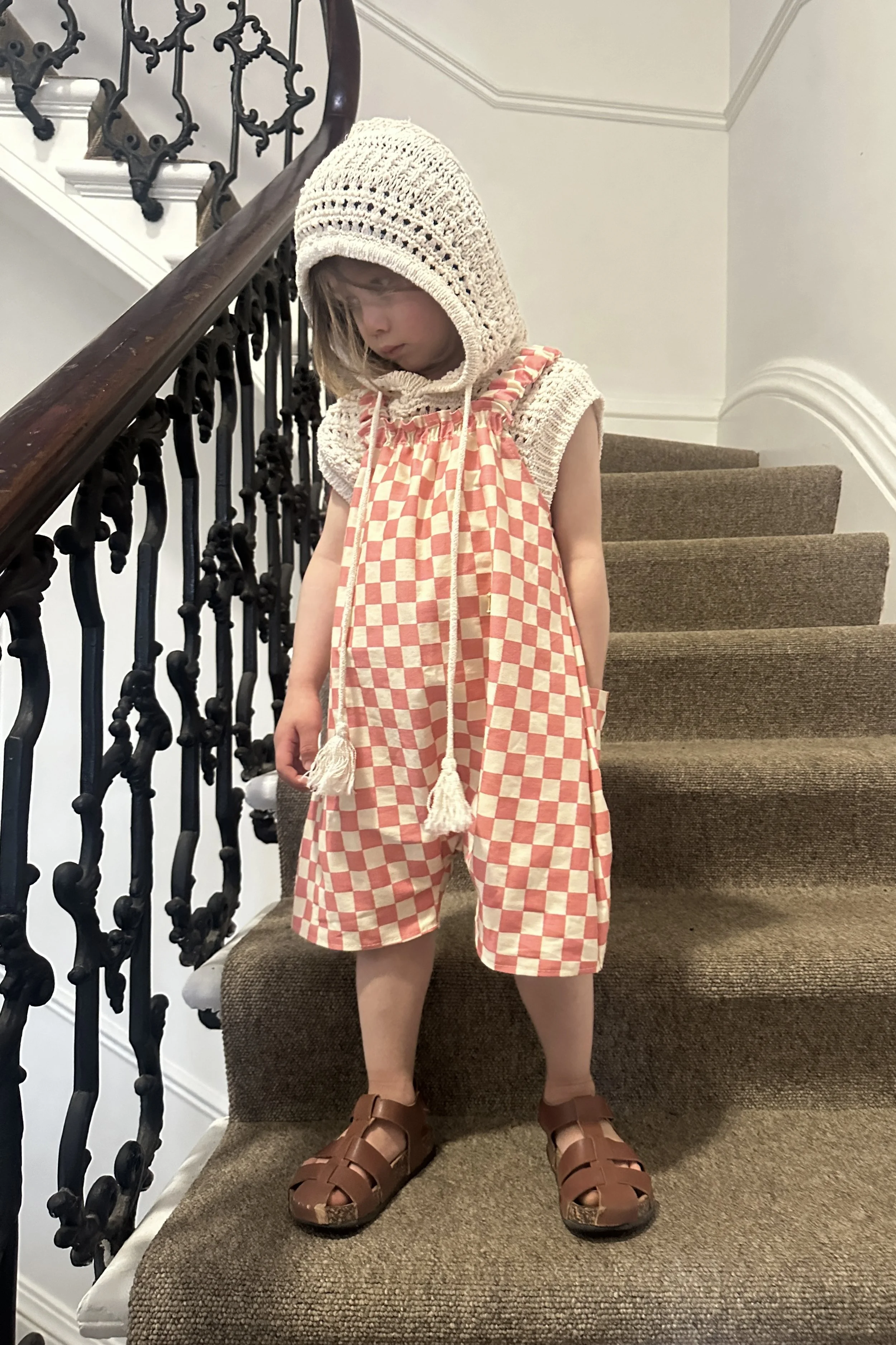 child wearing organic cotton canvas pullover short dungarees in pink checkerboard colour, layered over crochet short sleeves hoodie top, brown sandals, standing on staircase