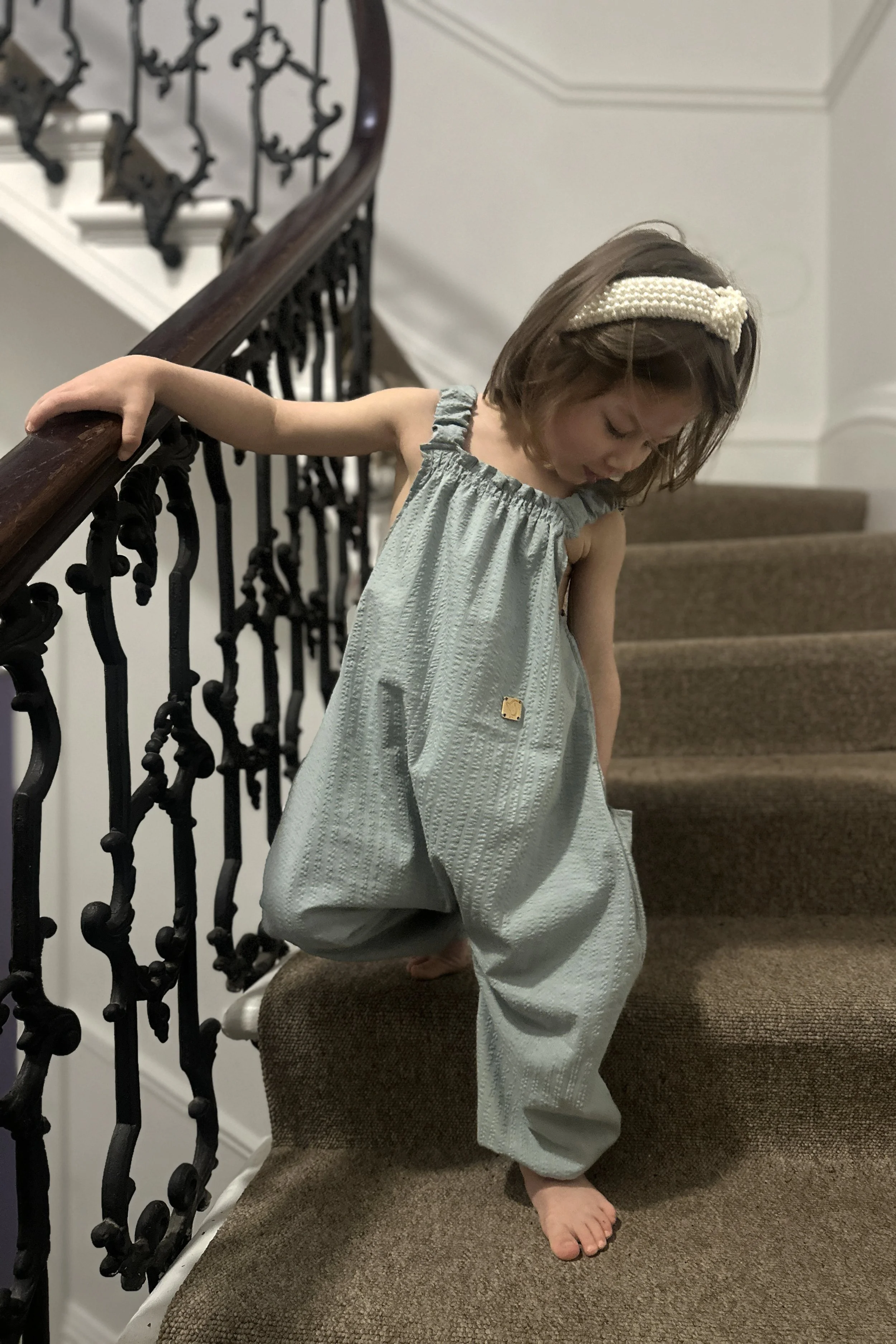 Child wearing organic cotton seersucker Duck Egg Blue pullover dungarees, pearl headband, standing on stairs, highlighting pocket at the back