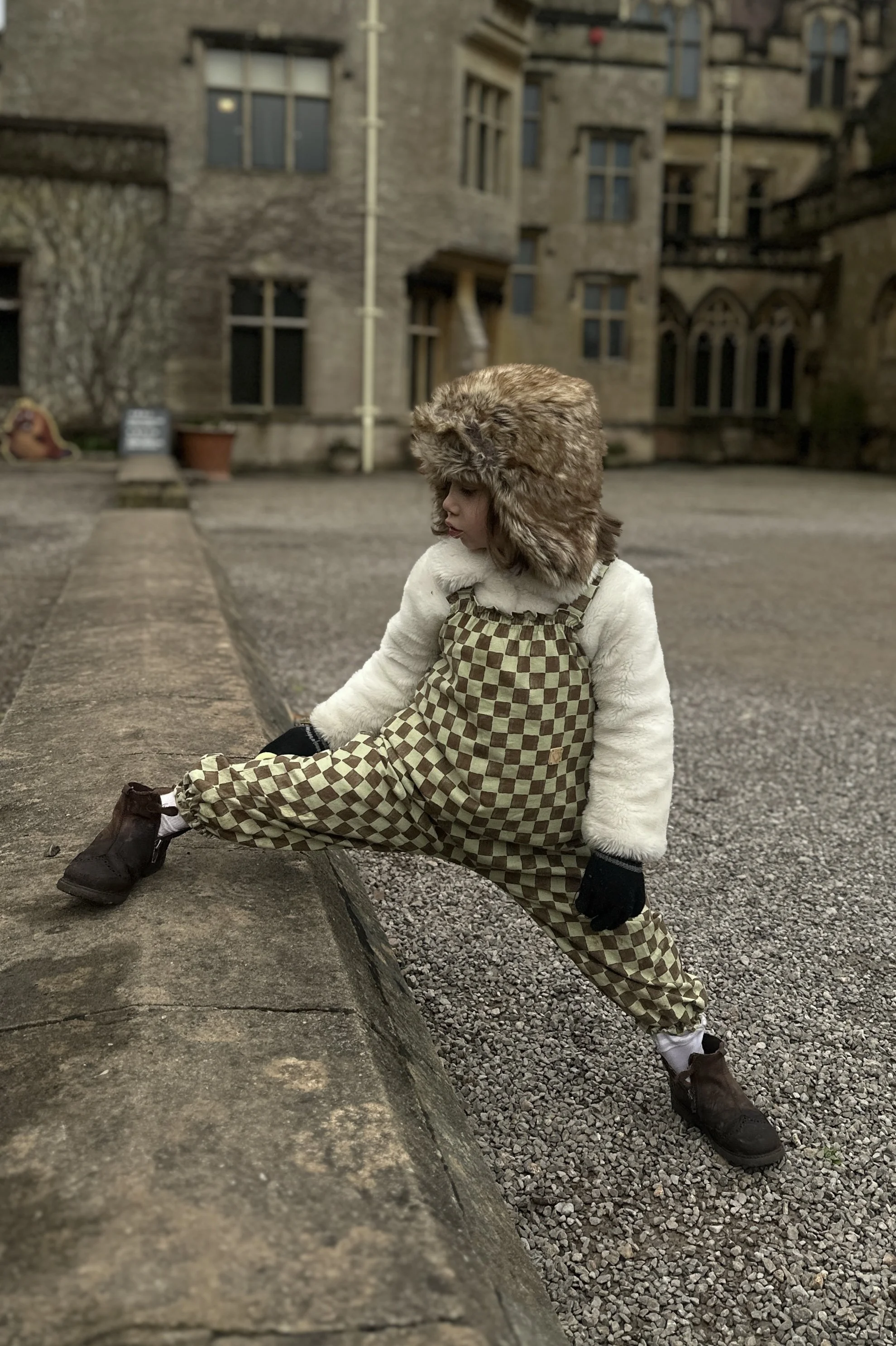 child wearing organic cotton checkerboard pullover dungarees in sage green and brown checks, faux fur trapper hat, brown boots, standing in front a tyntesfiled house