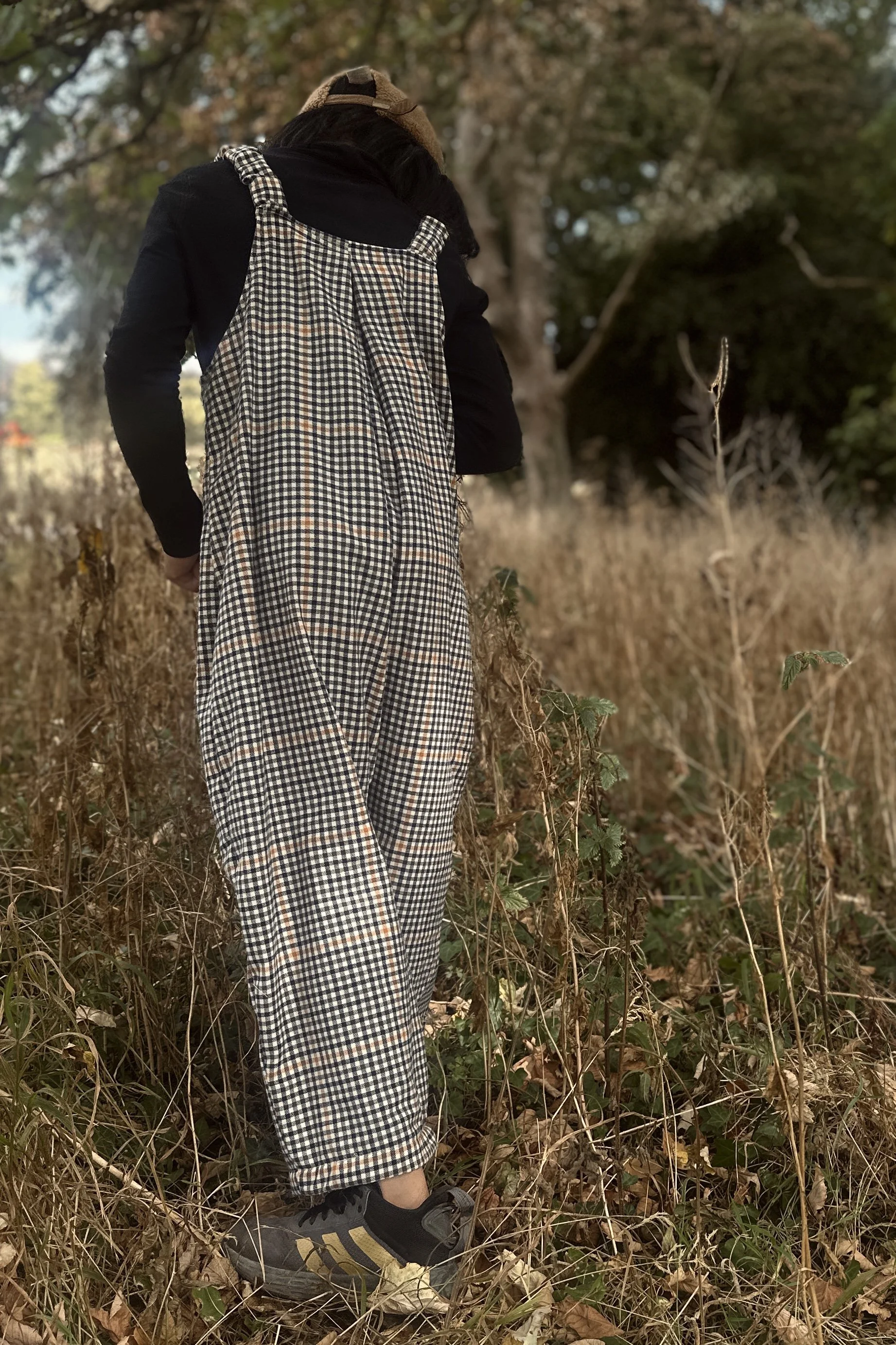 rear of adult wearing organic cotton retro brown check dungarees layered with black top, walking in dried grass