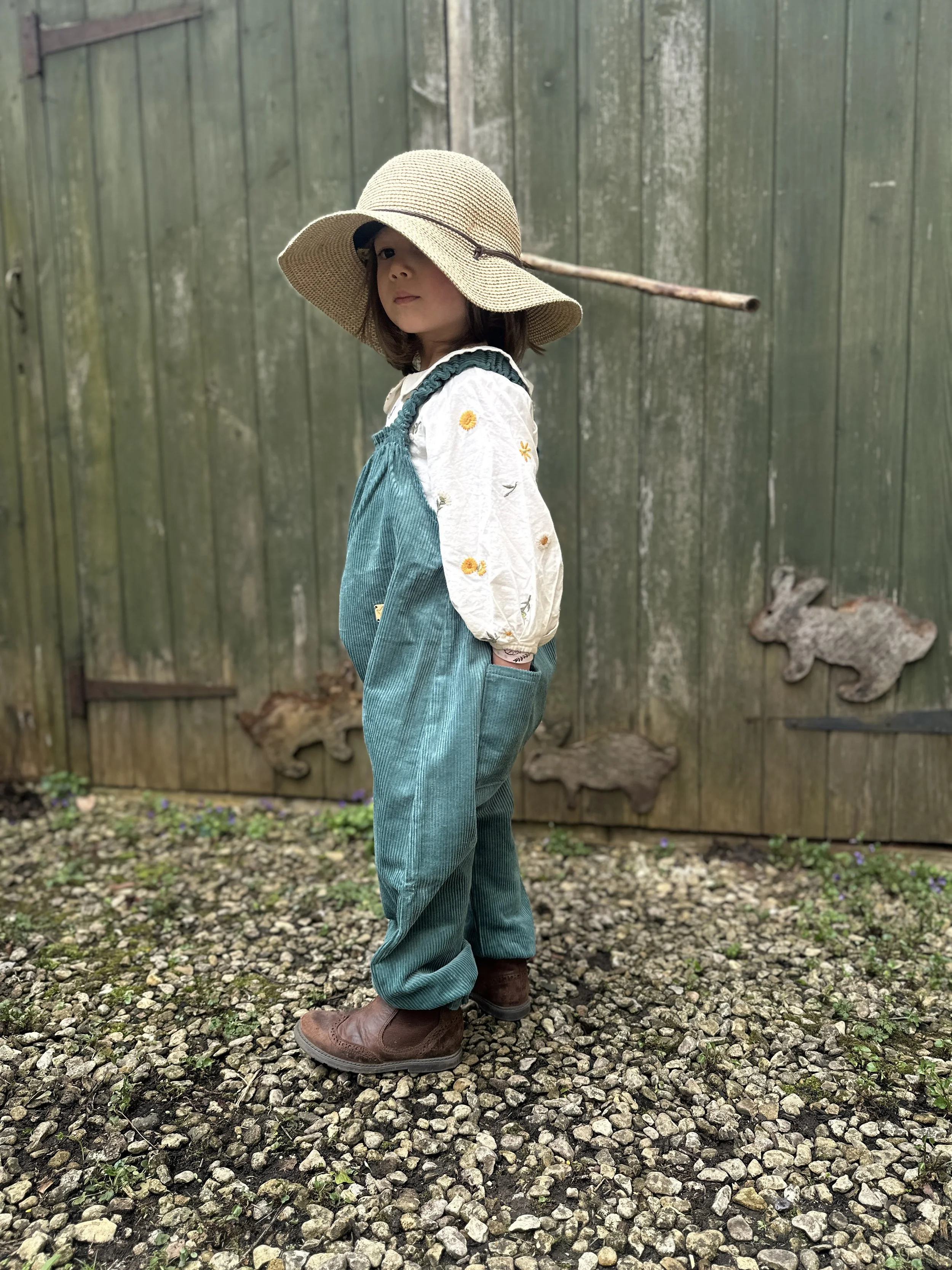 side view of child wearing organic cotton corduroy teal green pullover dungarees, brown boots, sun hat, standing in front of wooden shed