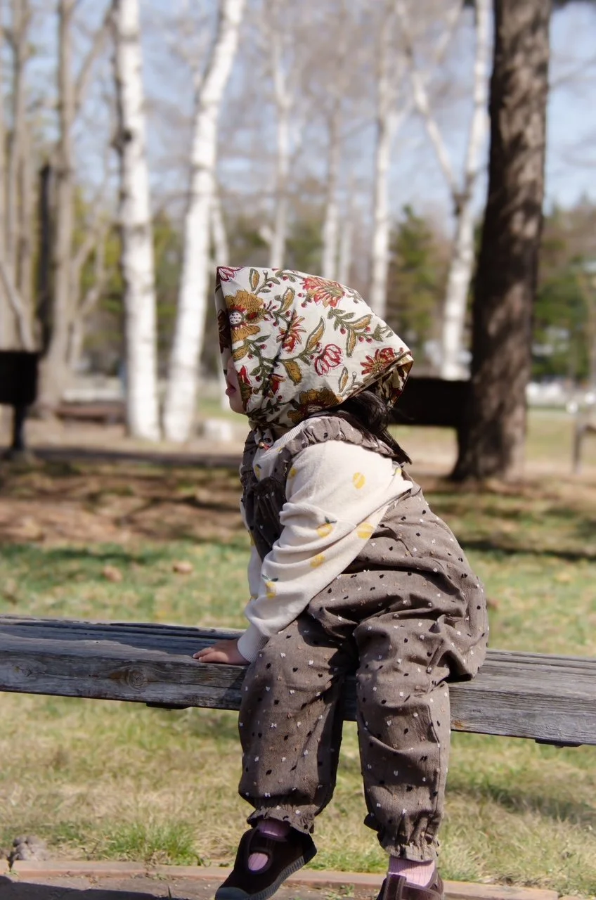 child wearing organic cotton speckled cocoa dungarees, floral handkerchief, sitting on a bench in the wood
