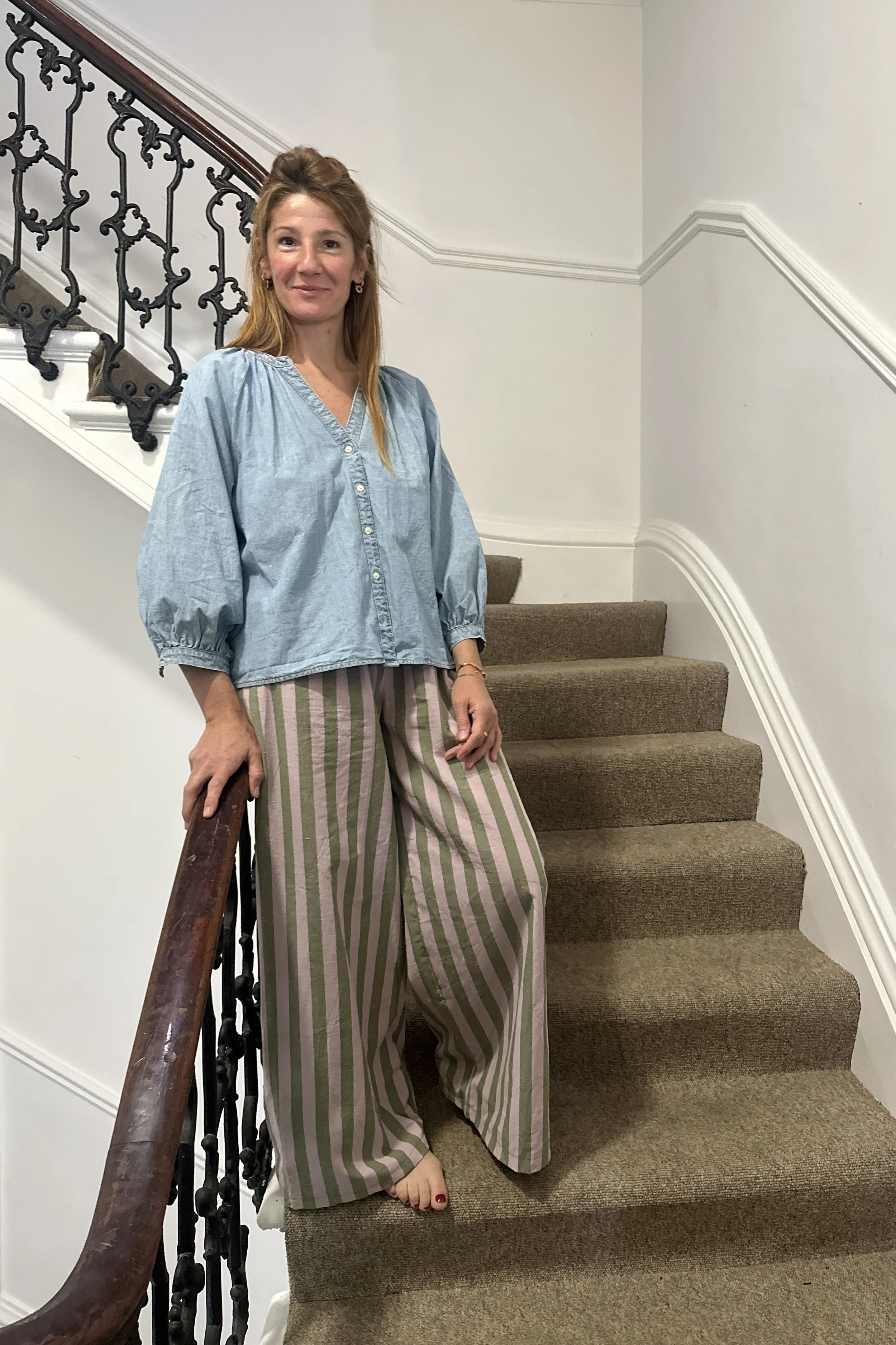 mum wearing organic cotton linen wide leg trousers stripes olive green and lilac, with denim light blue blouse standing on the stairs
