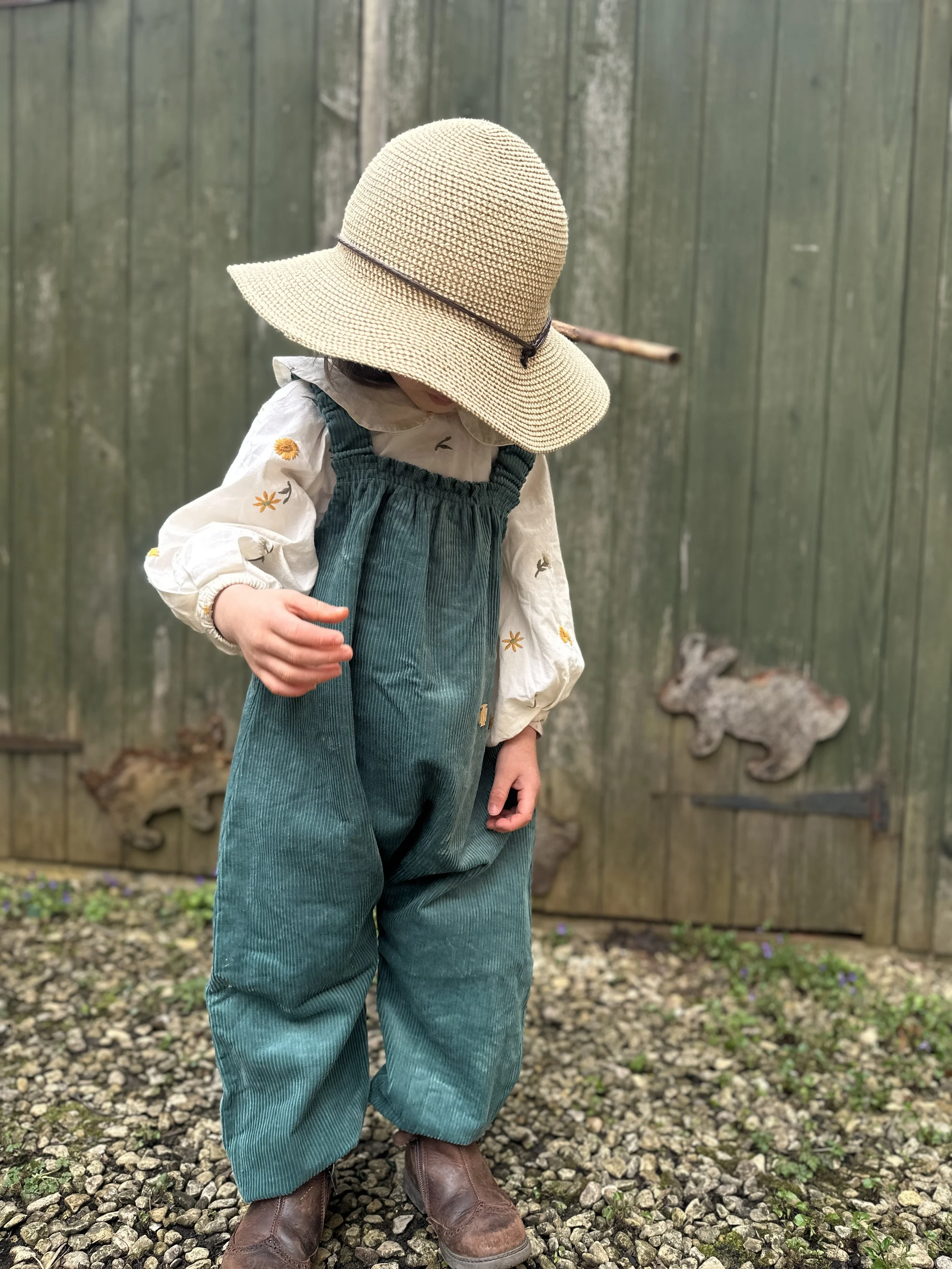 child wearing organic cotton corduroy teal green pullover dungarees, brown boots, sun hat, standing in front of wooden shed