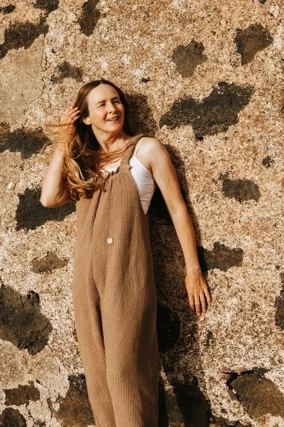 woman wearing organic cotton waffle pullover dungarees beige colour over a white t shirt, standing againt a cliff wall on the sea side