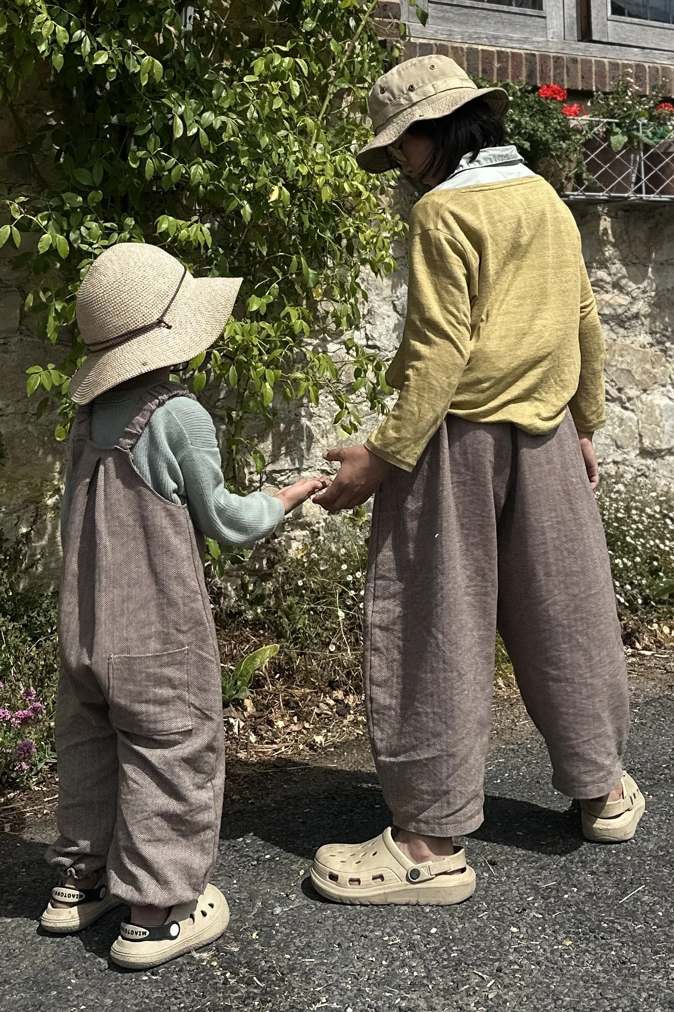adult wearing organic cotton herringbone brown barrel leg trousers full length, standing with a child wearing wearing organic cotton herringbone brown dungarees
