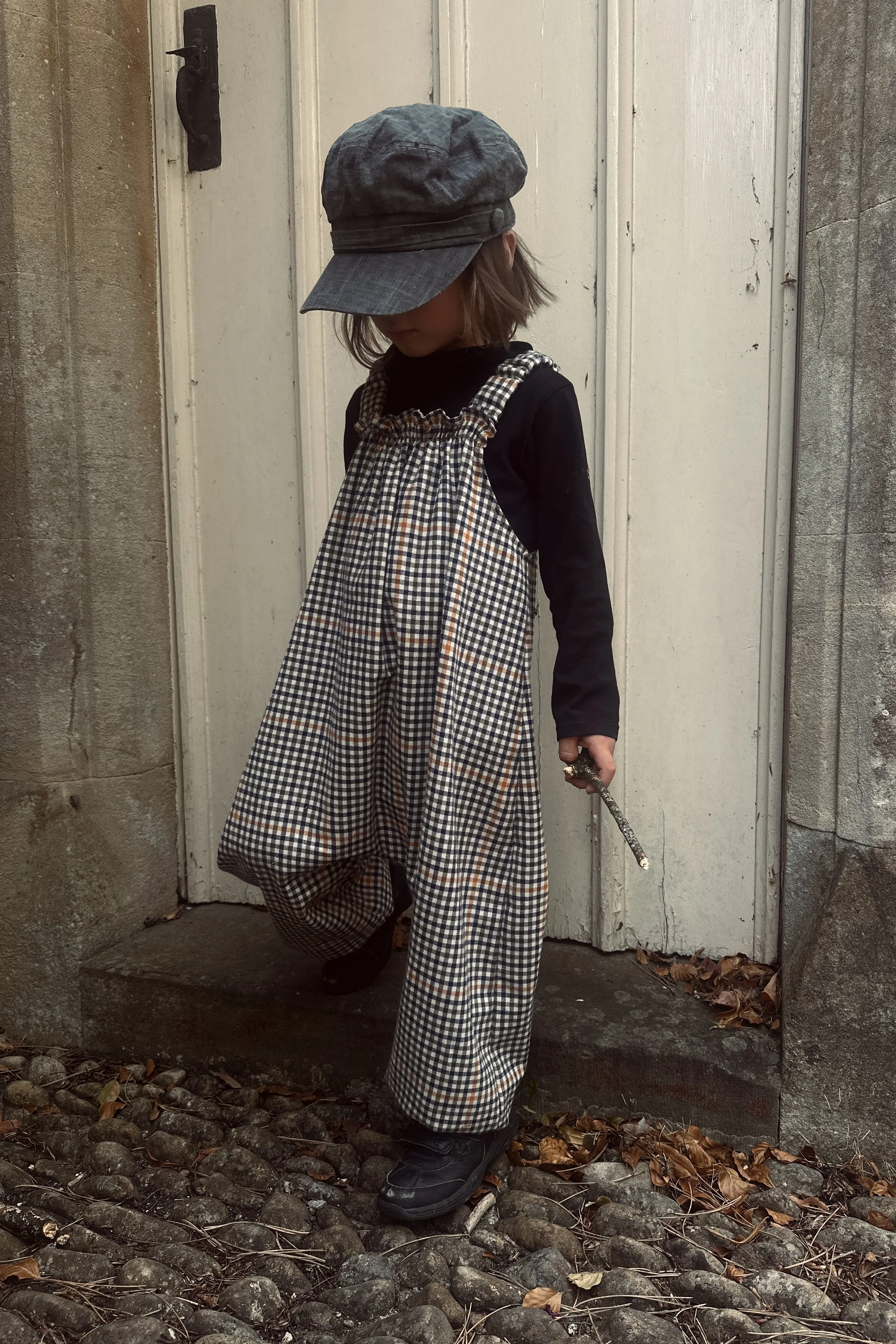 child wearing organic cotton retro brown check dungarees layered with black top, grey hat, standing in front of a white door
