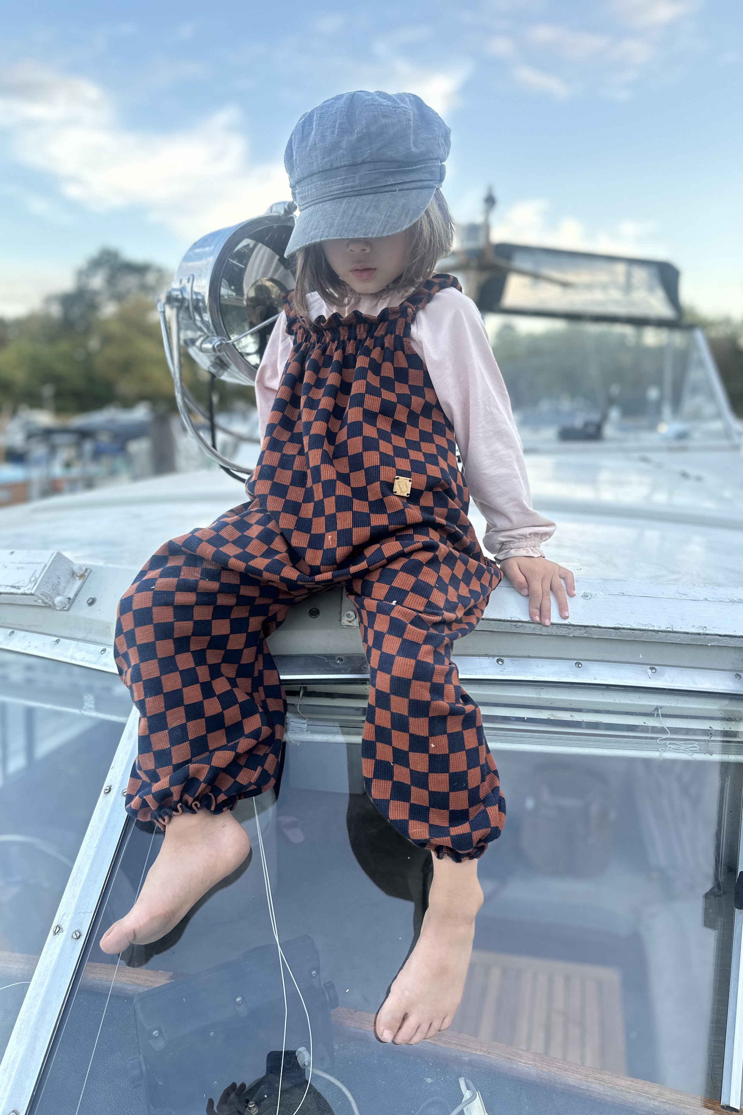child wearing pullover dungarees in checkerboard navy and rust colour, sitting on a boat at marina