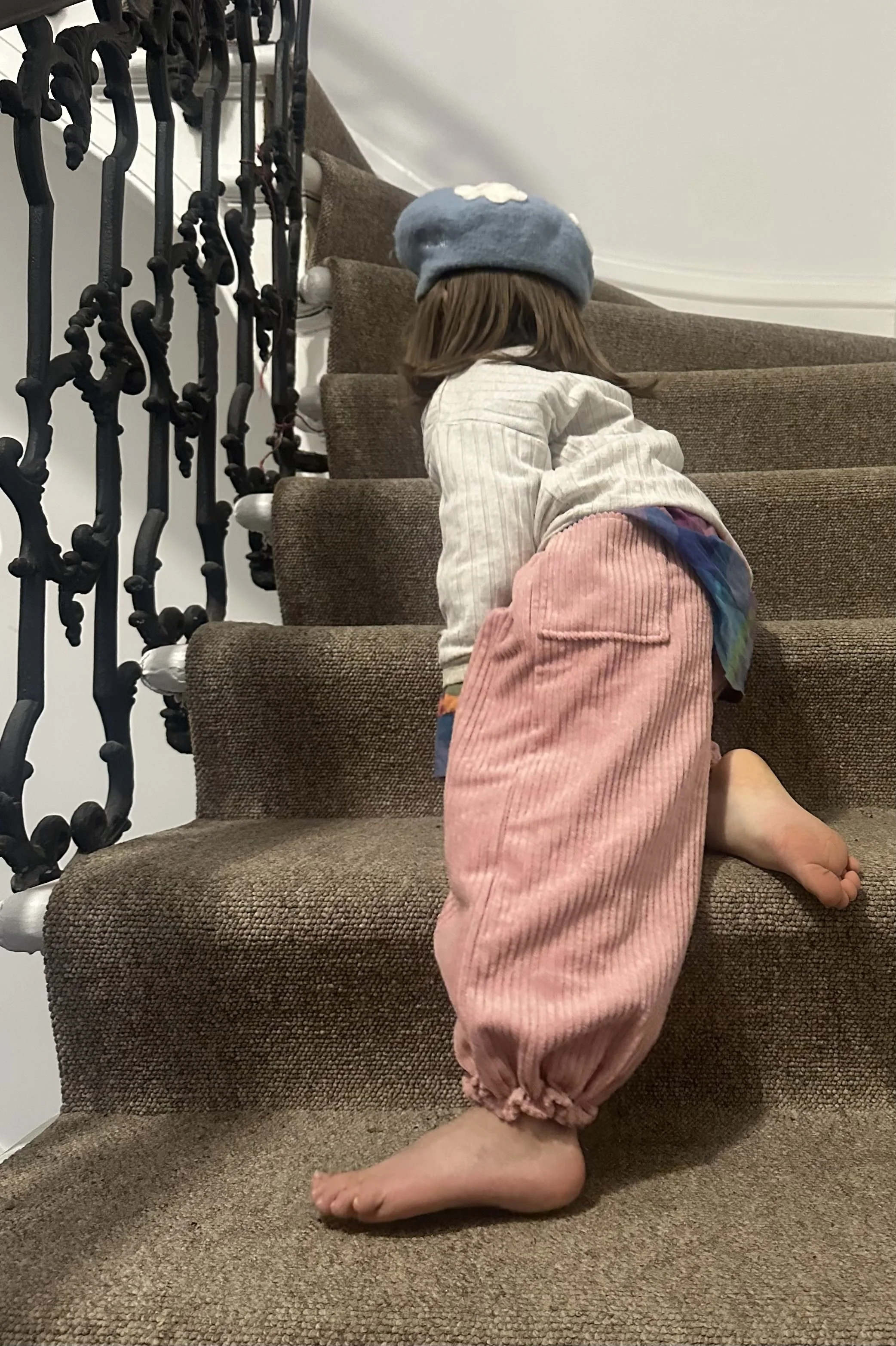 rear of child wearing organic cotton corduroy pink trousers barrel shape baggy fit oversized elastic hem, with white top, walking up staircase, highlighting pocket at back
