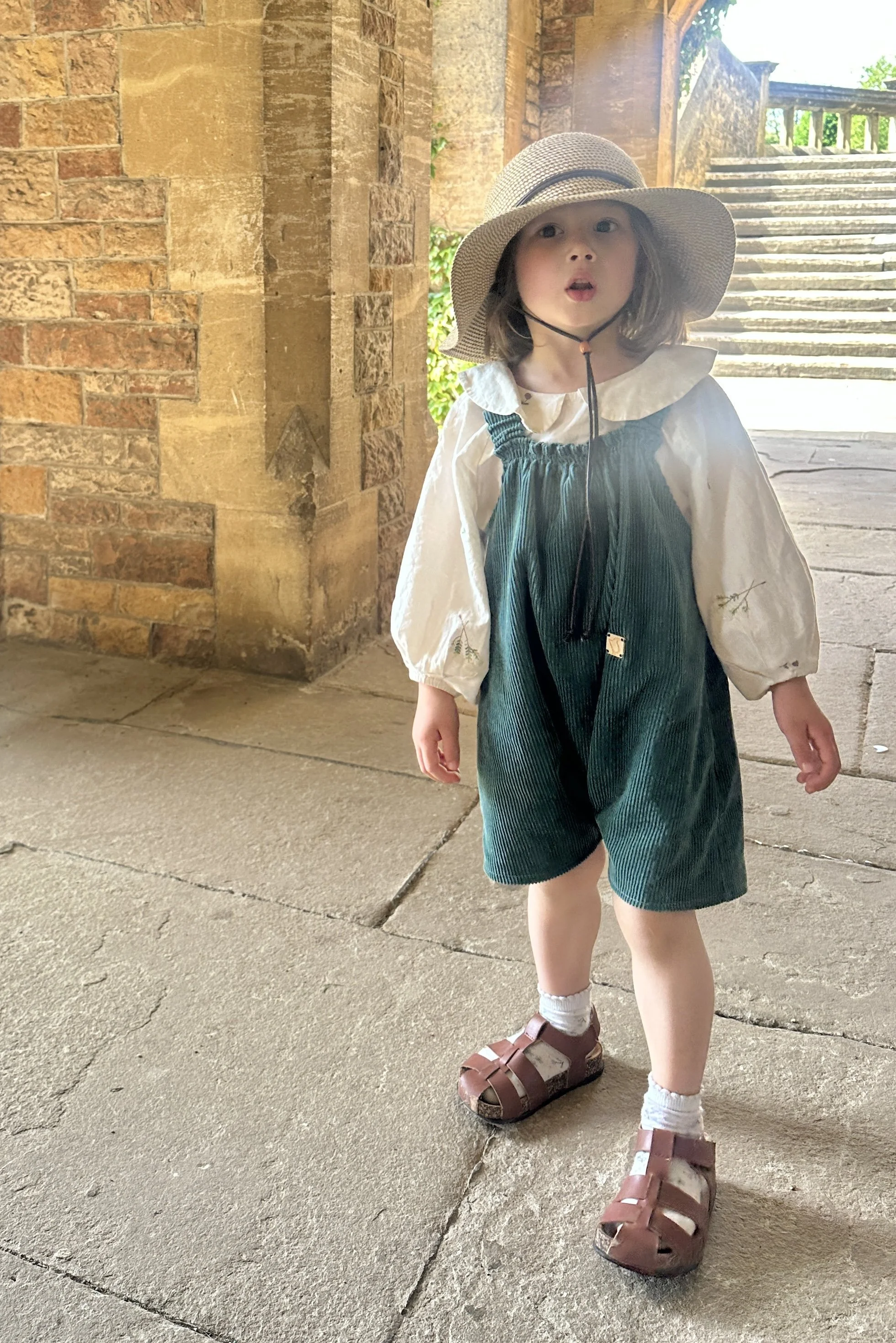 Child wearing teal green organic cotton corduroy short dungarees styled with a white embroidered blouse, sun hat, and brown sandals, standing outdoors under a stone archway in warm sunlight