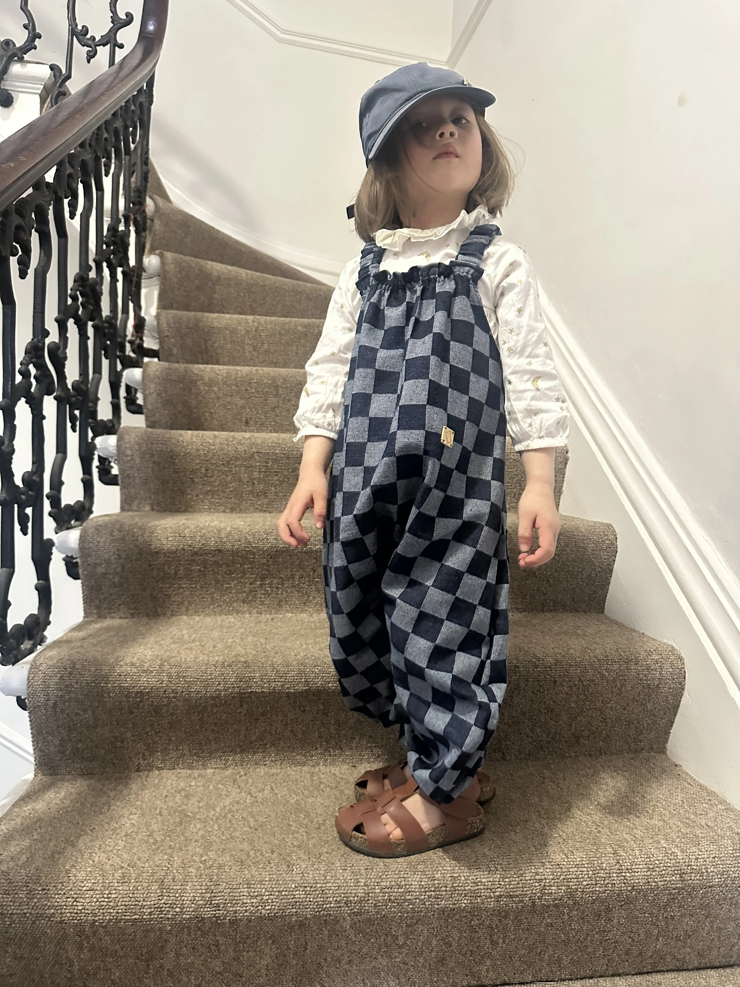 child wearing organic cotton checkerboard denim blue dungarees, brown sandals, denim cap, standing on staircase