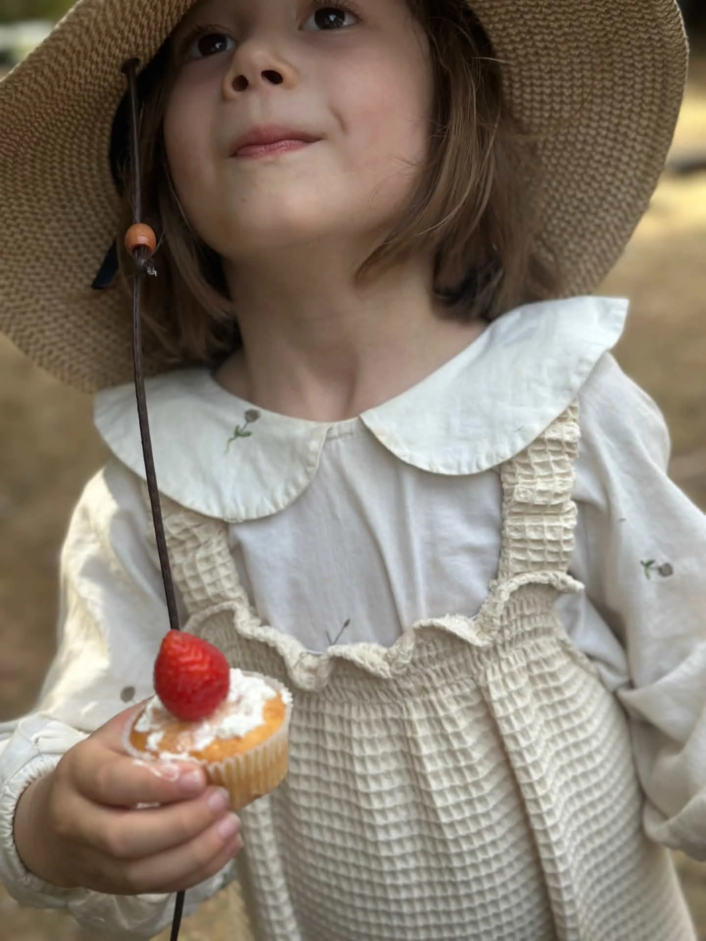 close up child wearing organic cotton waffle natural white dungarees layered with white blouse, sun hat, holding a cupcake with strawberry