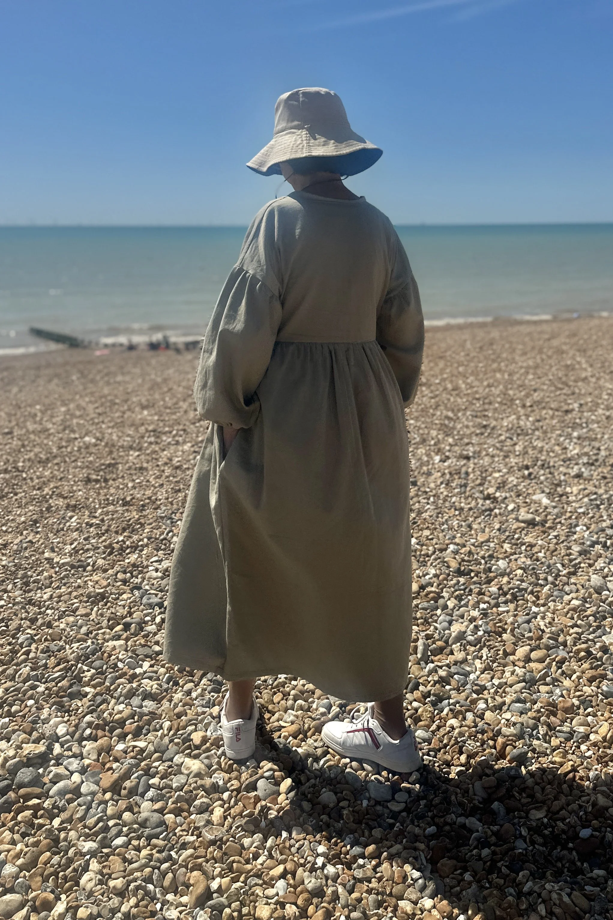 rear view of adult wearing organic cotton denim light sage maxi dress, white trainers, beige sunhat, walking on the beach