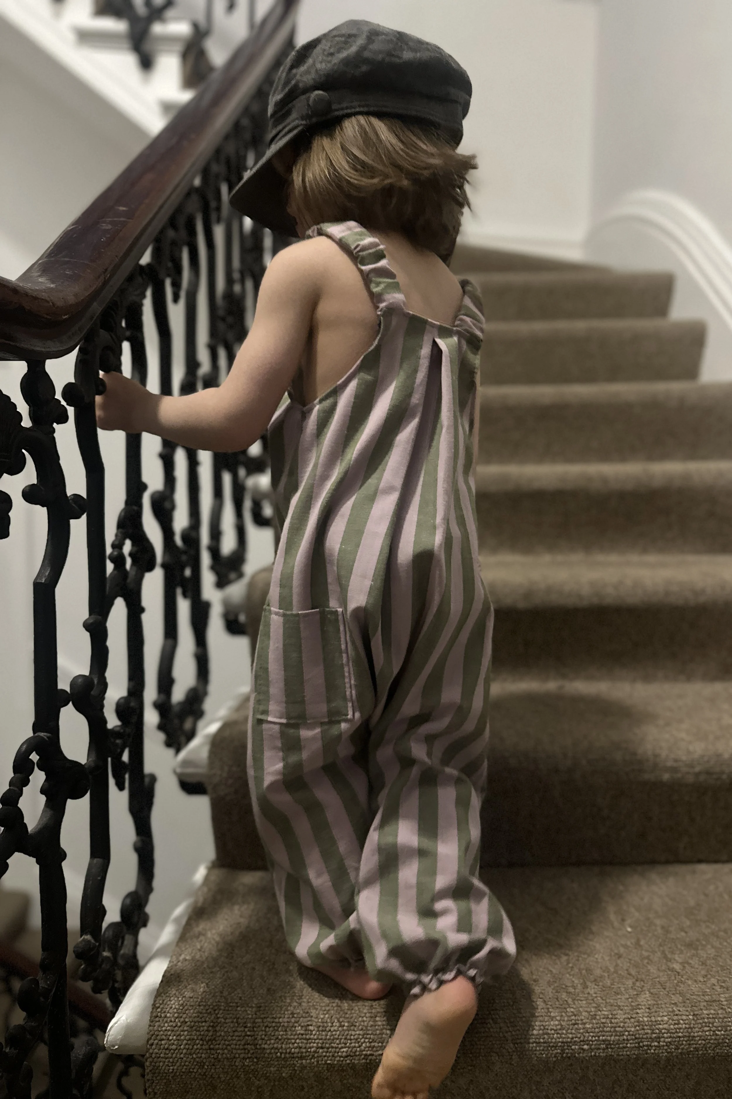 back view of Child wearing organic cotton and linen dungarees in wide stripe olive green and lilac, and a grey hat, standing on staircase (Copy)