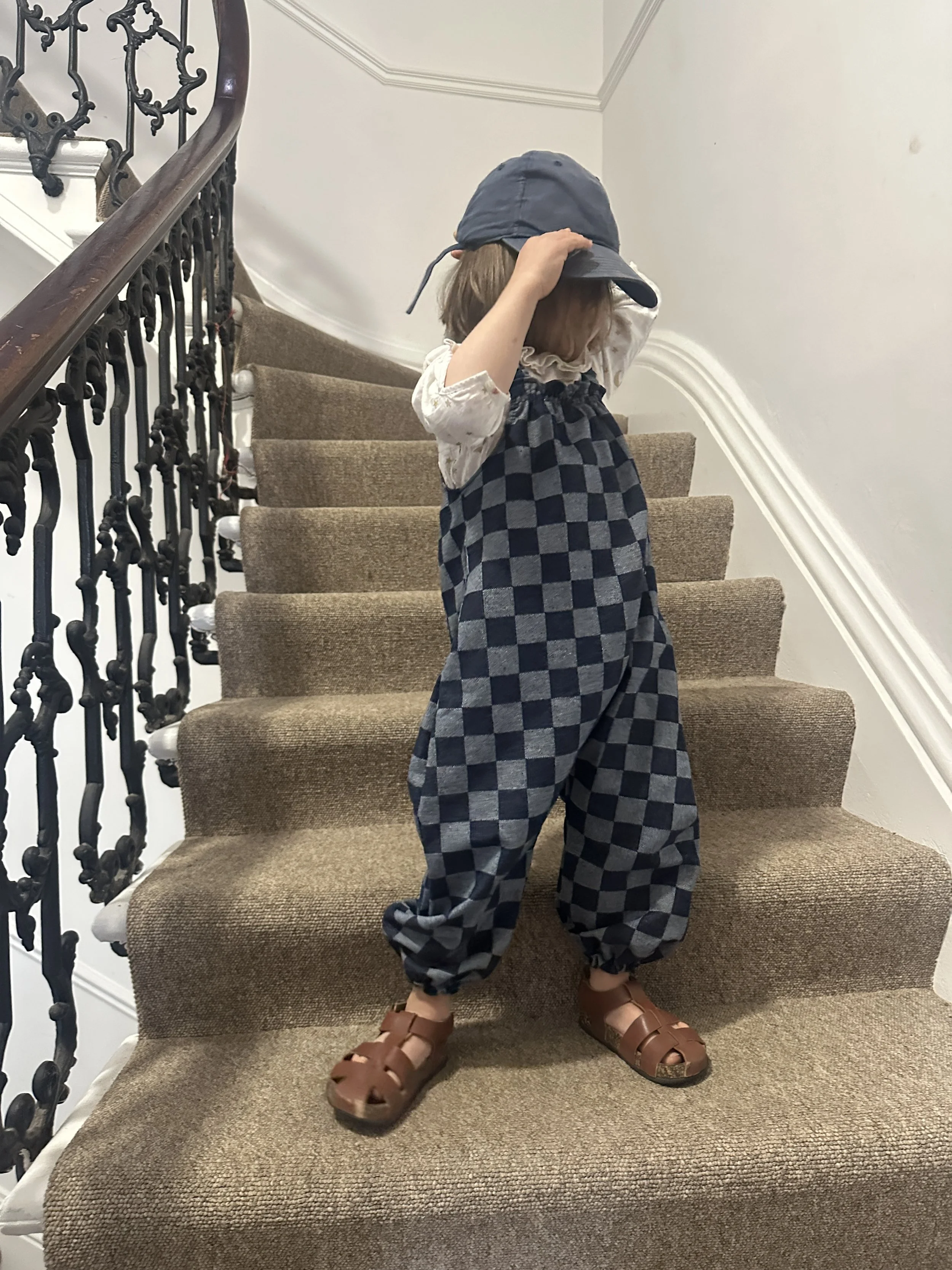child wearing organic cotton checkerboard denim blue dungarees, brown sandals, denim cap, standing on staircase