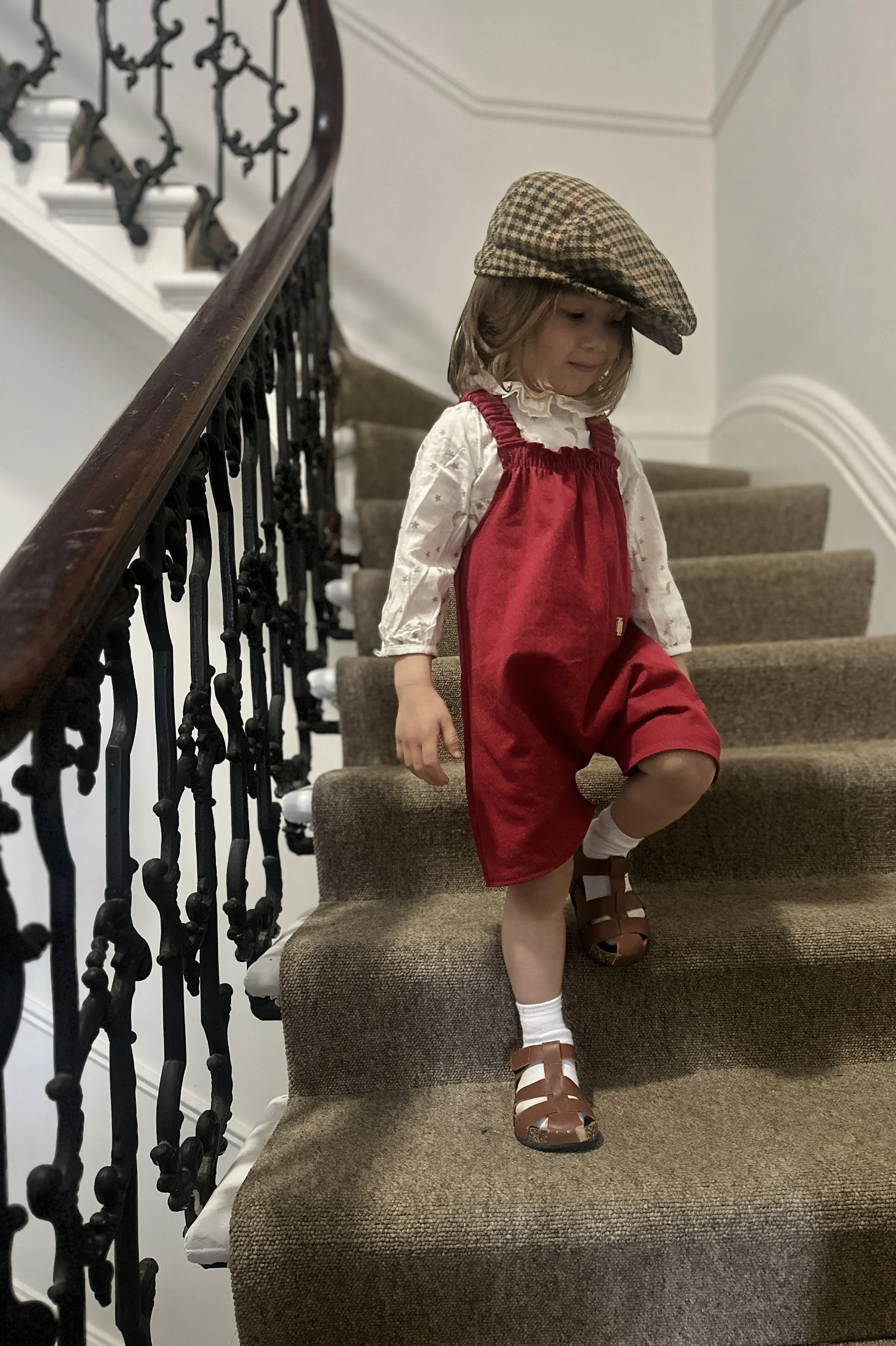 child wearing organic cotton denim red short dungarees over white blouse with golden stars with brown sandals tartan hat, standing on staircase