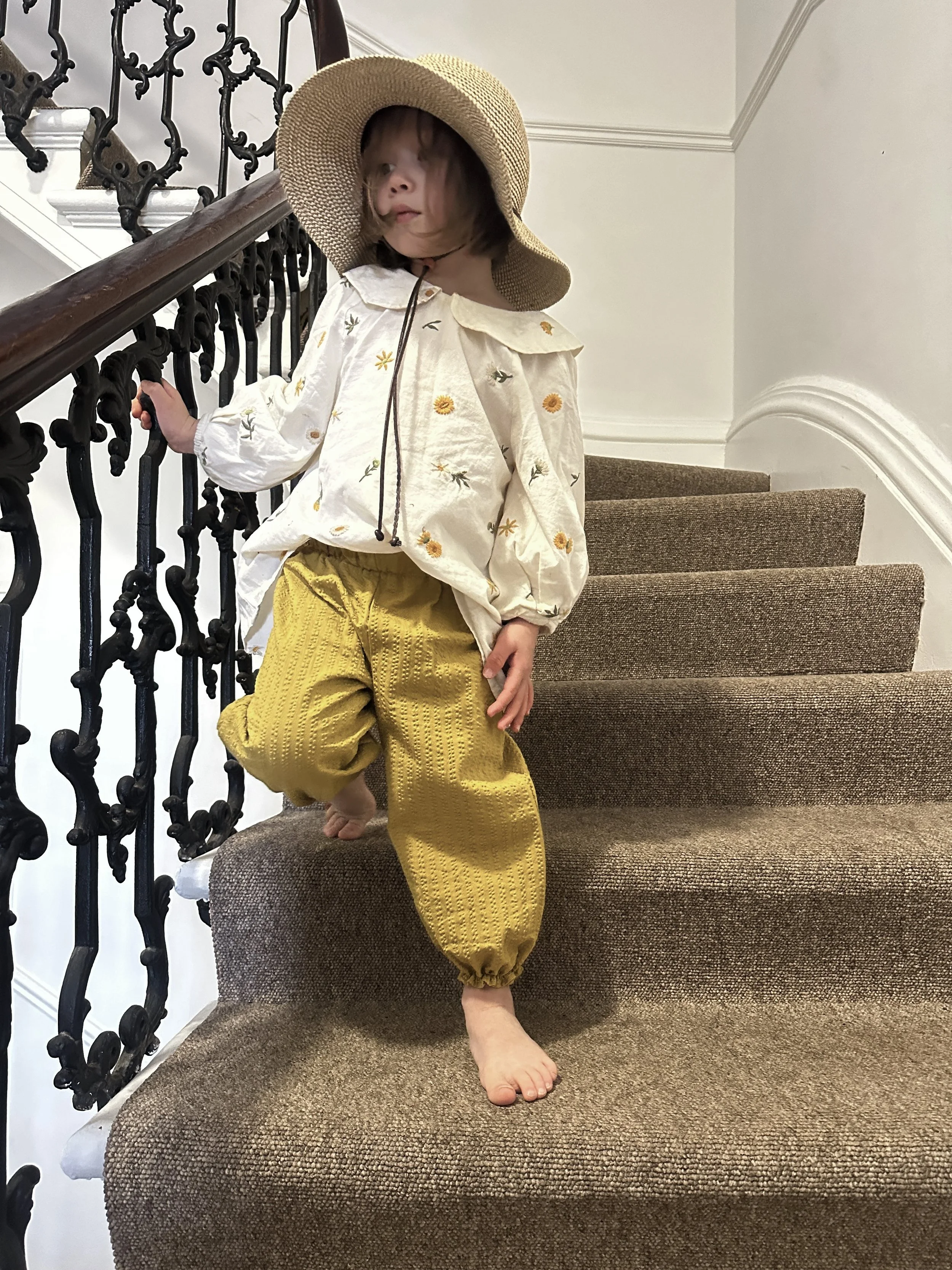 child wearing organic cotton seersucker barrel trousers mustard colour, paired with white blouse with Peter Pan collar and embroidered flowers, sun hat