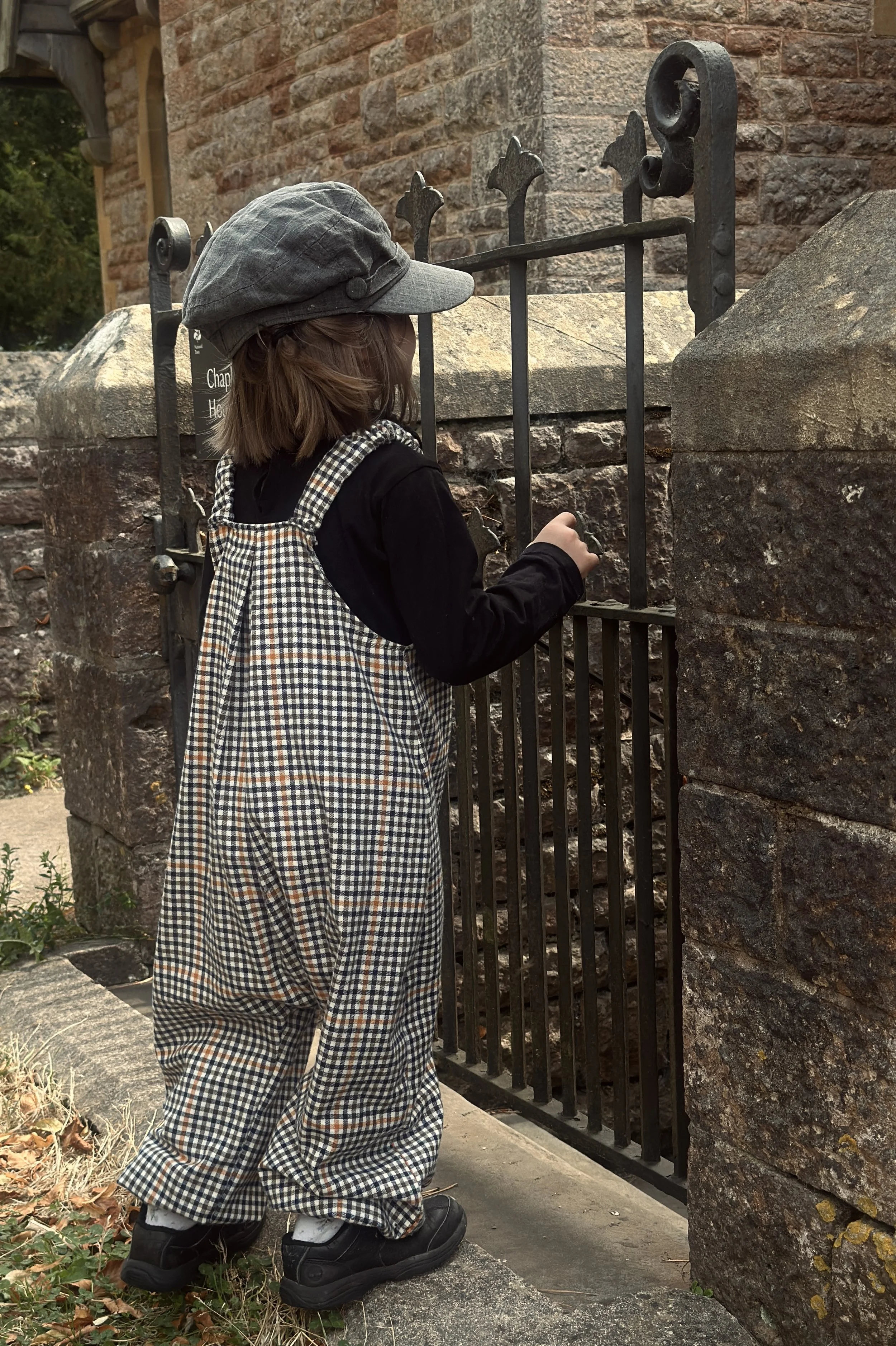 rear of child wearing organic cotton retro brown check dungarees layered with black top, grey hat,