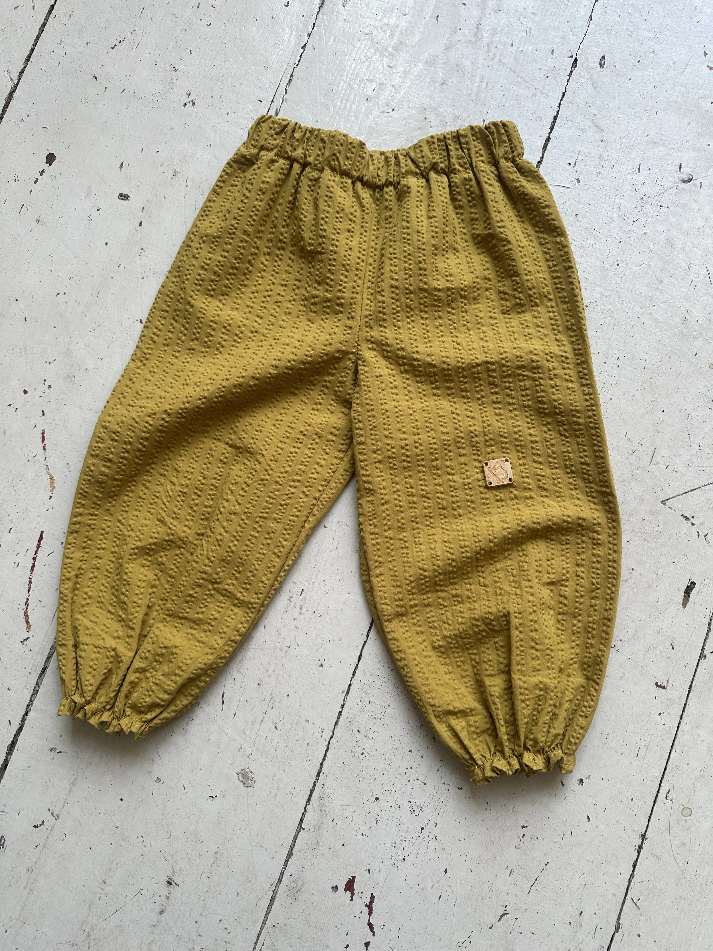 flat lay front of organic cotton seersucker barrel trousers mustard colour