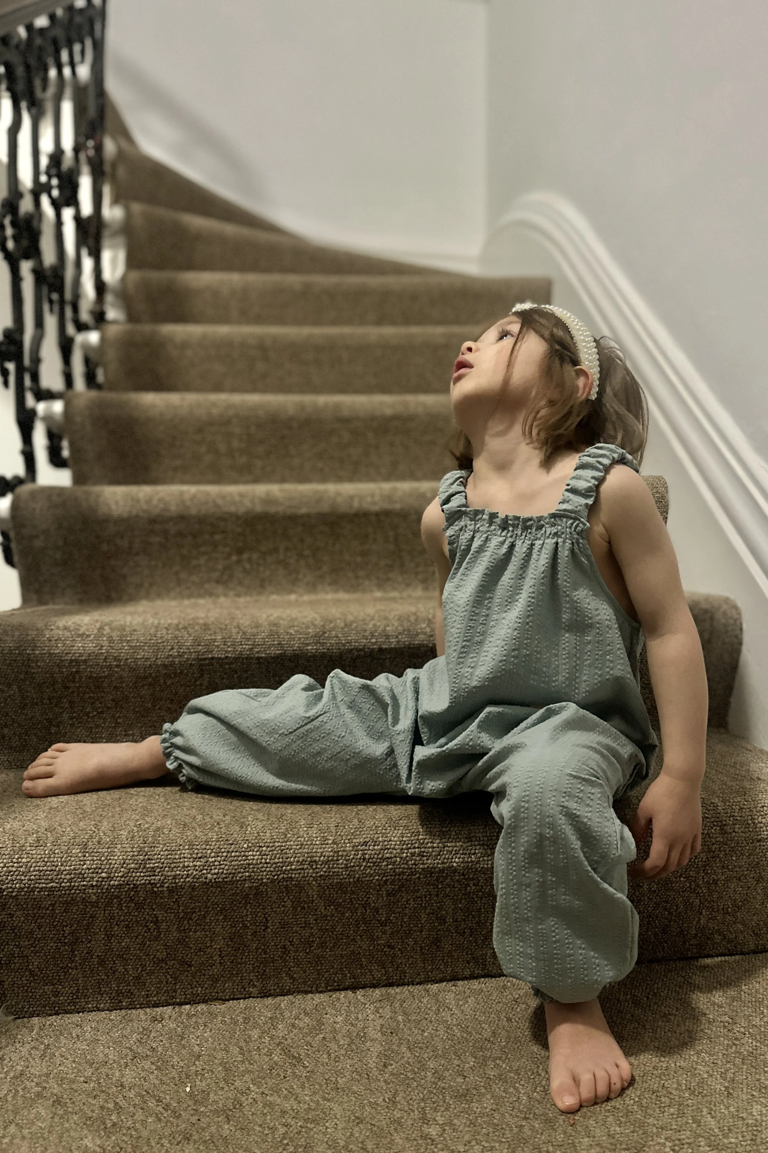 Child wearing organic cotton seersucker Duck Egg Blue pullover dungarees, pearl headband, sitting on stairs