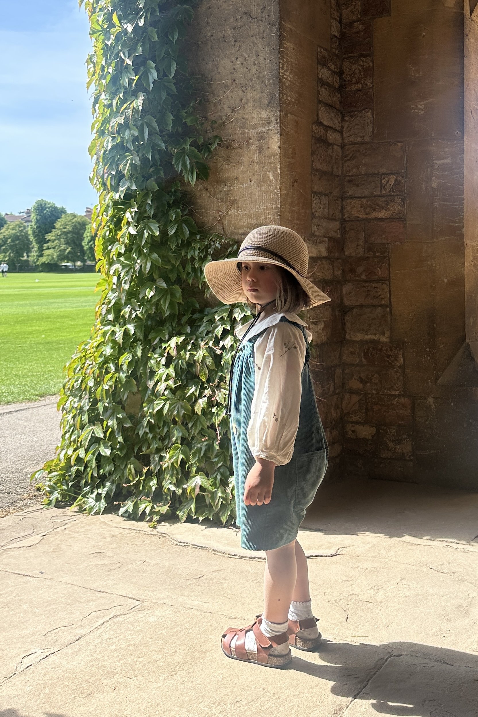 Child wearing teal green organic cotton corduroy short dungarees styled with a white embroidered blouse, sun hat, and brown sandals, standing outdoors under a stone archway in warm sunlight