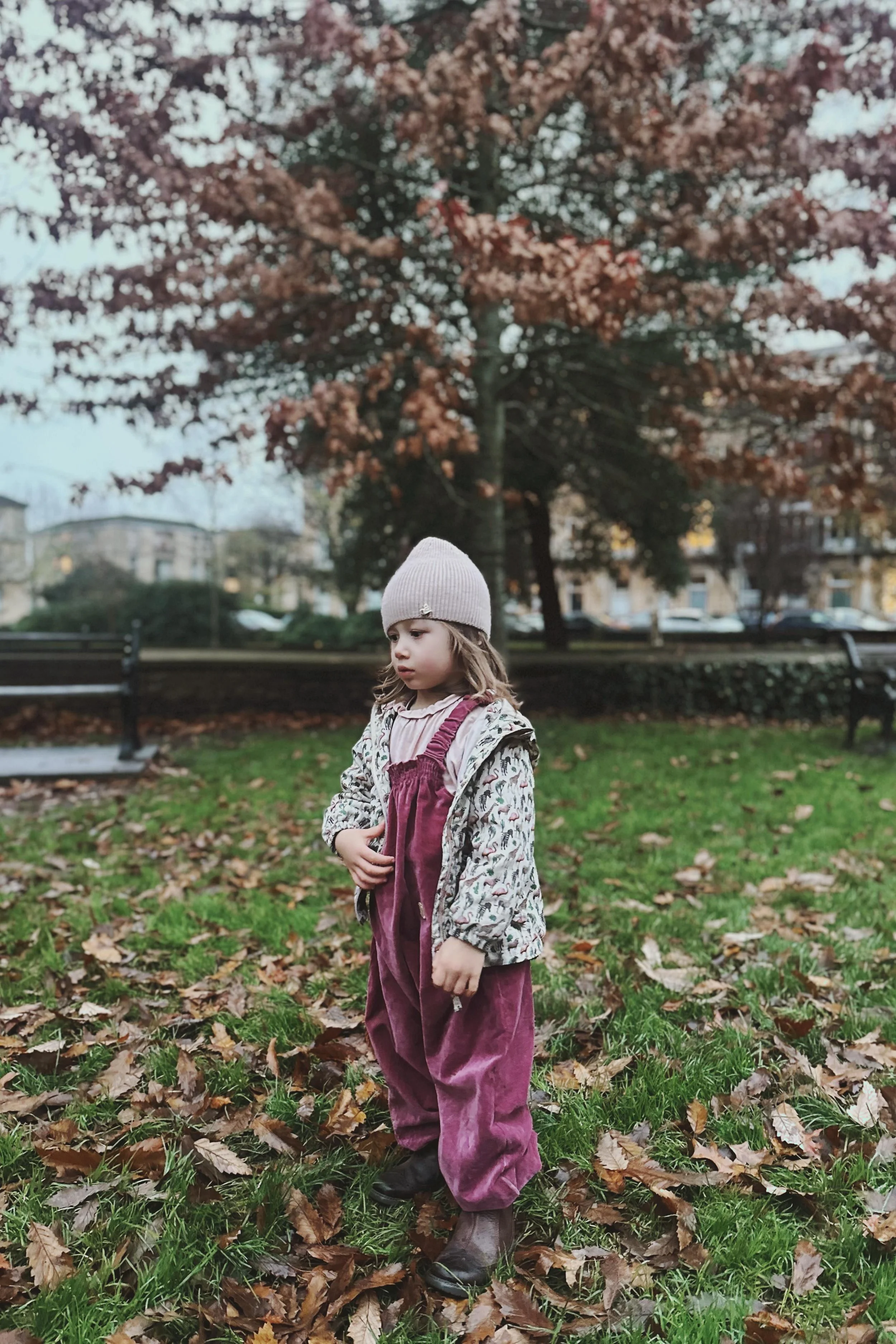 Child wearing dark rose pink organic velvet pullover dungarees with raincoat, pink woolly hat, and brown boots, running in a park in the evening