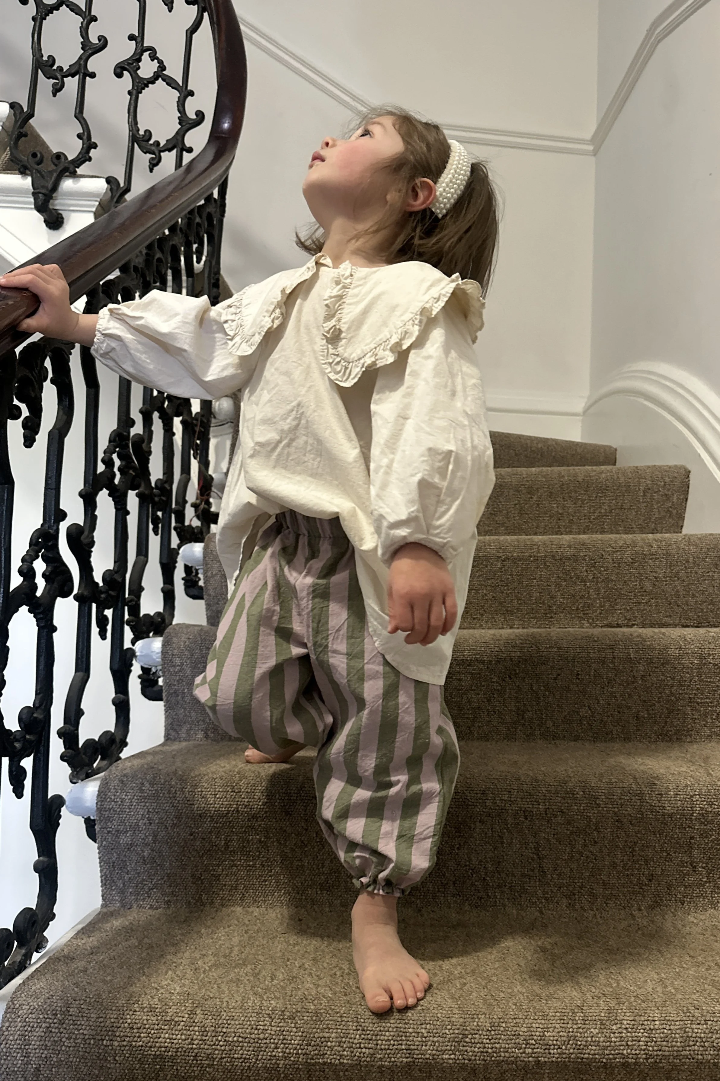 child wearing organic cotton wide stripe barrel trousers in olive green and lilac colour, paired with cream blouse with large frill collars, brown sandals, pearl hairband, standing on staircase