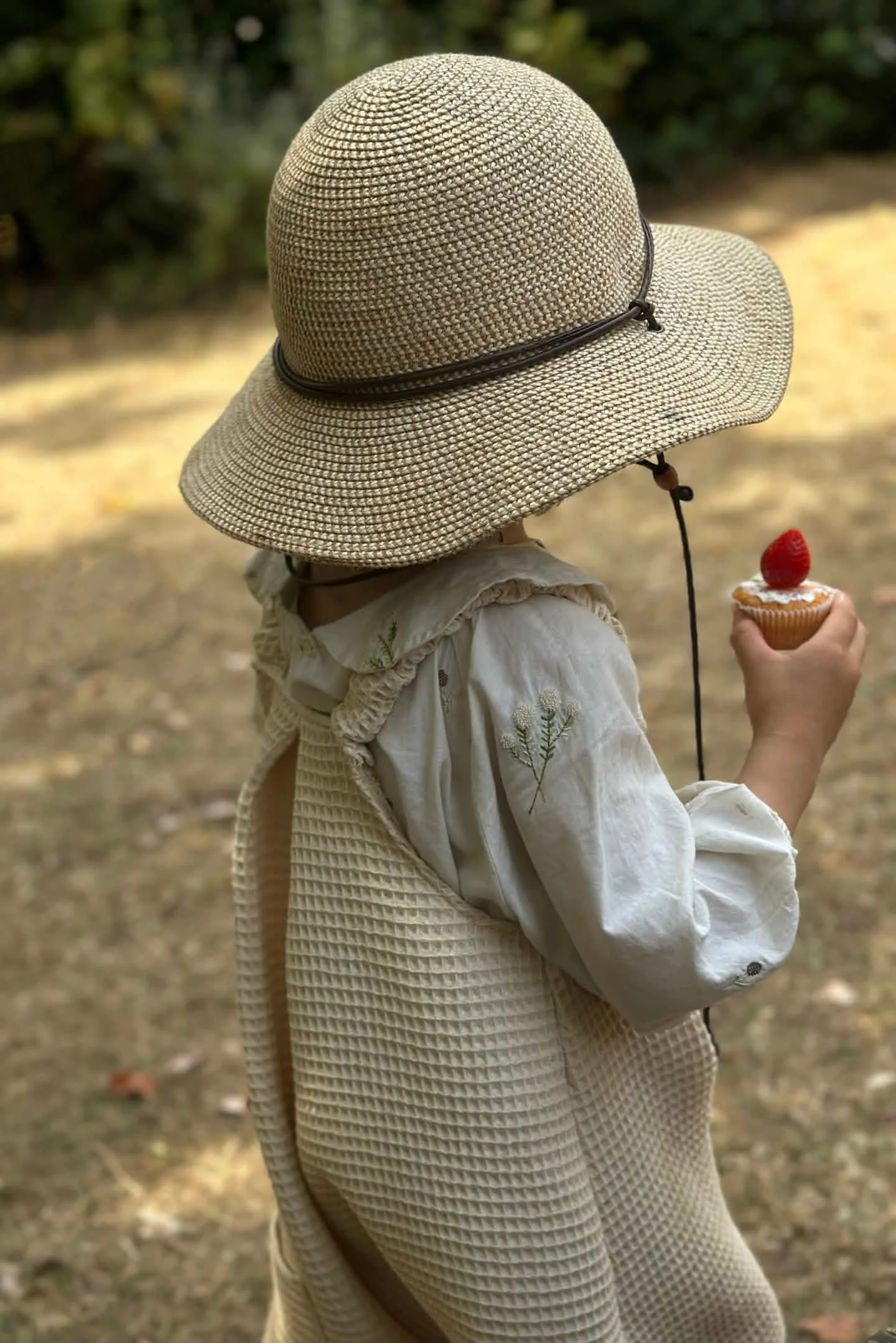 child wearing organic cotton waffle natural white dungarees layered with white blouse, sun hat, holding a cupcake with strawberry